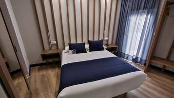 A modern hotel room with a large bed, wooden panel walls, a bedside table, and a window with curtains.
