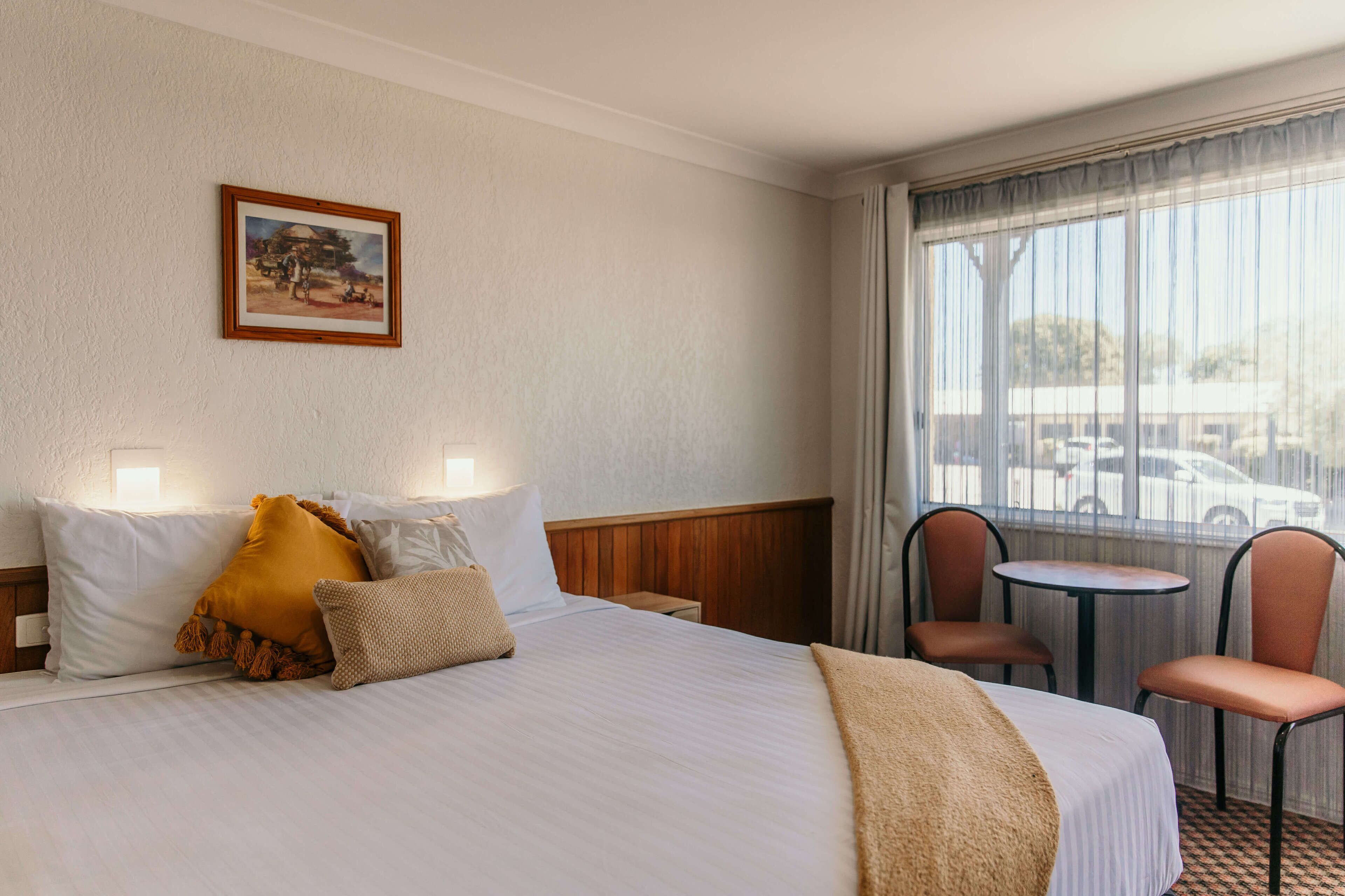 Habitación de hotel con cama, dos sillas, una mesita, un cuadro en la pared y ventana con vista a coches aparcados.