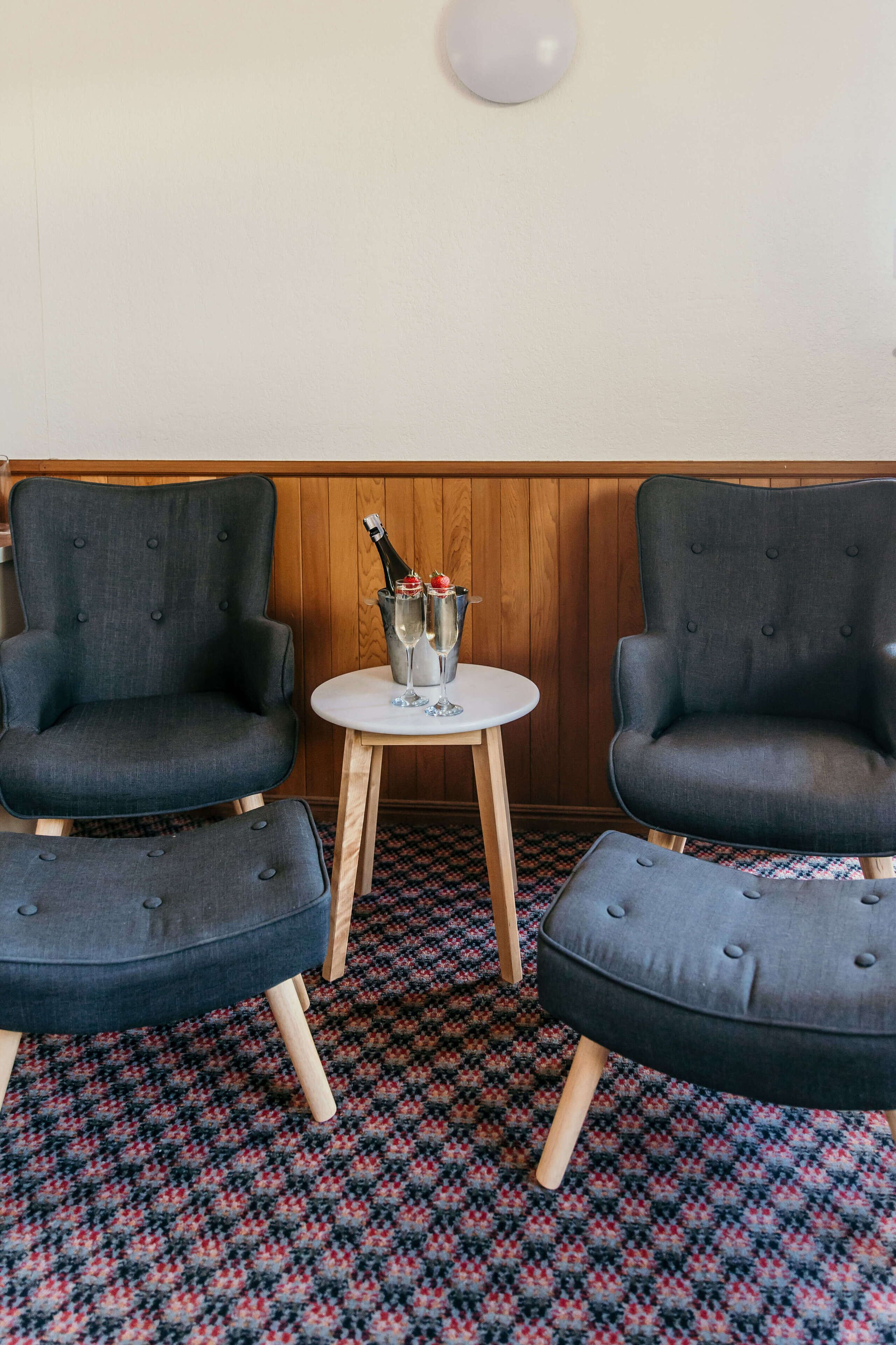 Two upholstered armchairs with wooden legs, a small round table with a bottle of champagne, and a patterned carpet.