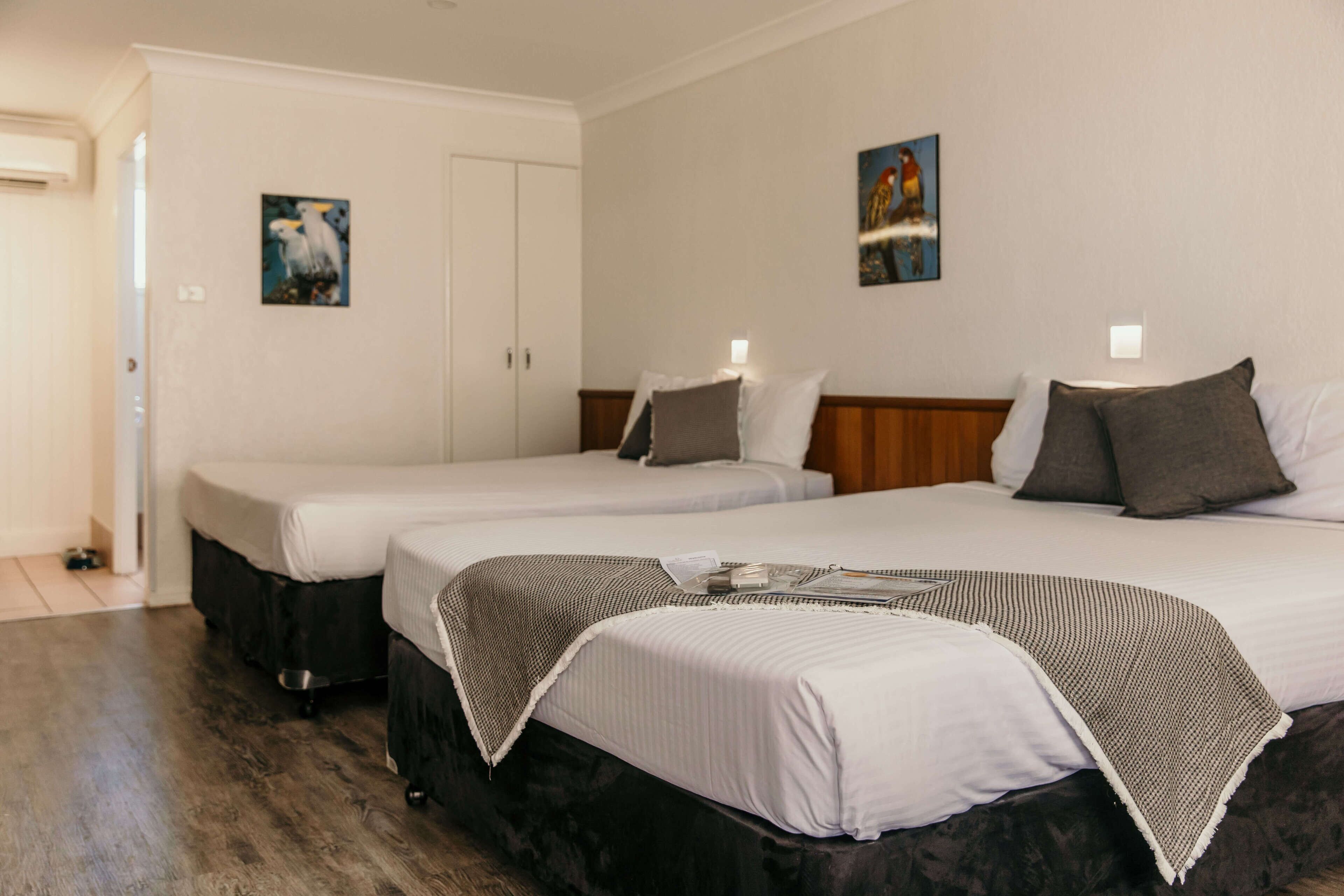A hotel room with two single beds, a large bed, and a wooden headboard. There are two paintings on the walls and a door leading to another room.