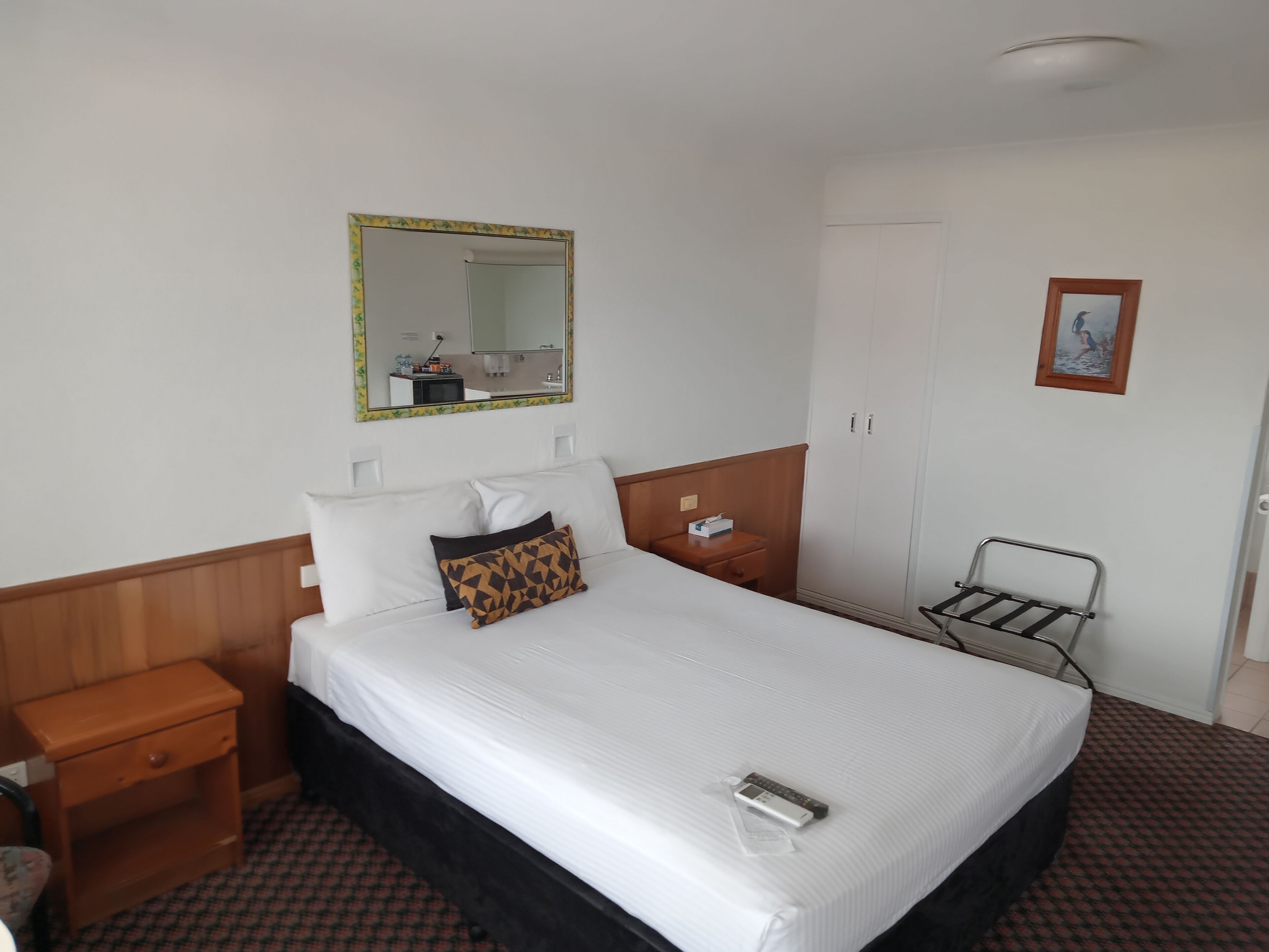 A hotel room with a bed, a bedside table, a nightstand, a mirror, a wardrobe, and a small shelf.
