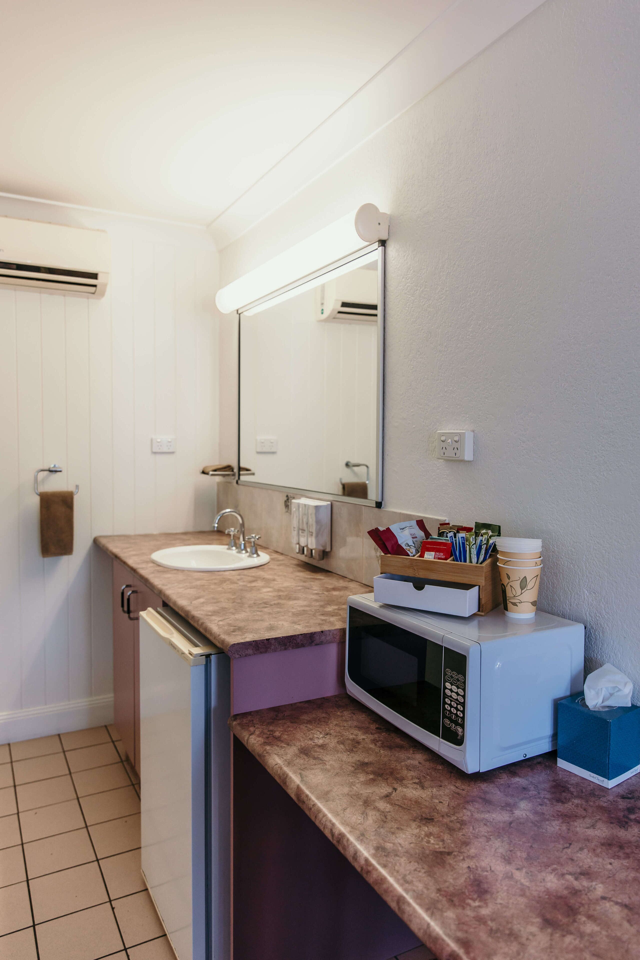 A bathroom with a sink, mirror, microwave, and a small storage area with toiletries.