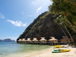 Overwater bungalows with thatched roofs, a beach, clear turquoise water, and a mountainous backdrop. - Miniloc Island Resort (El Nido)
