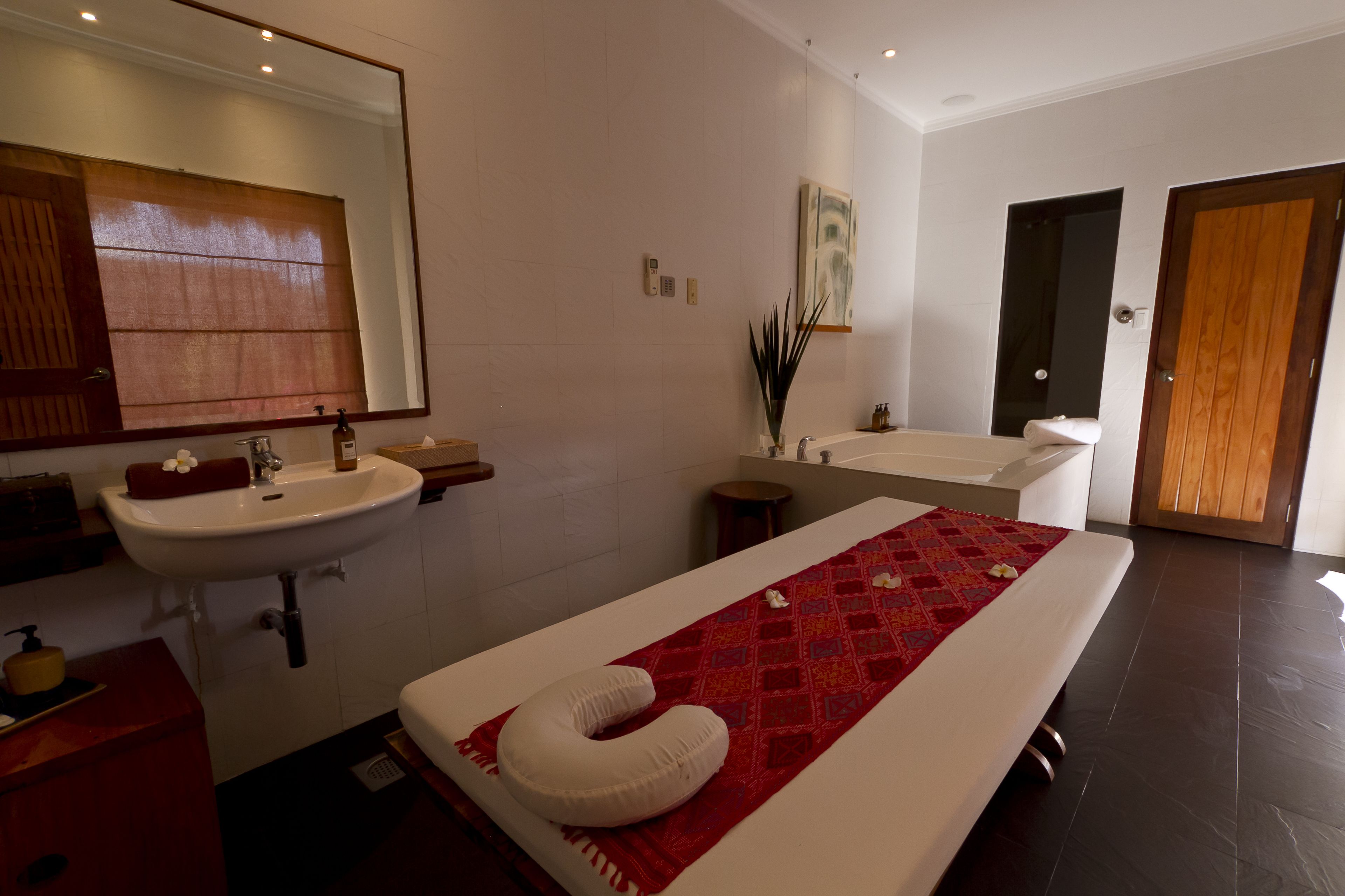 Un baño moderno con un espejo grande, dos lavamanos, una camilla de masaje con una toalla roja y una puerta de madera.