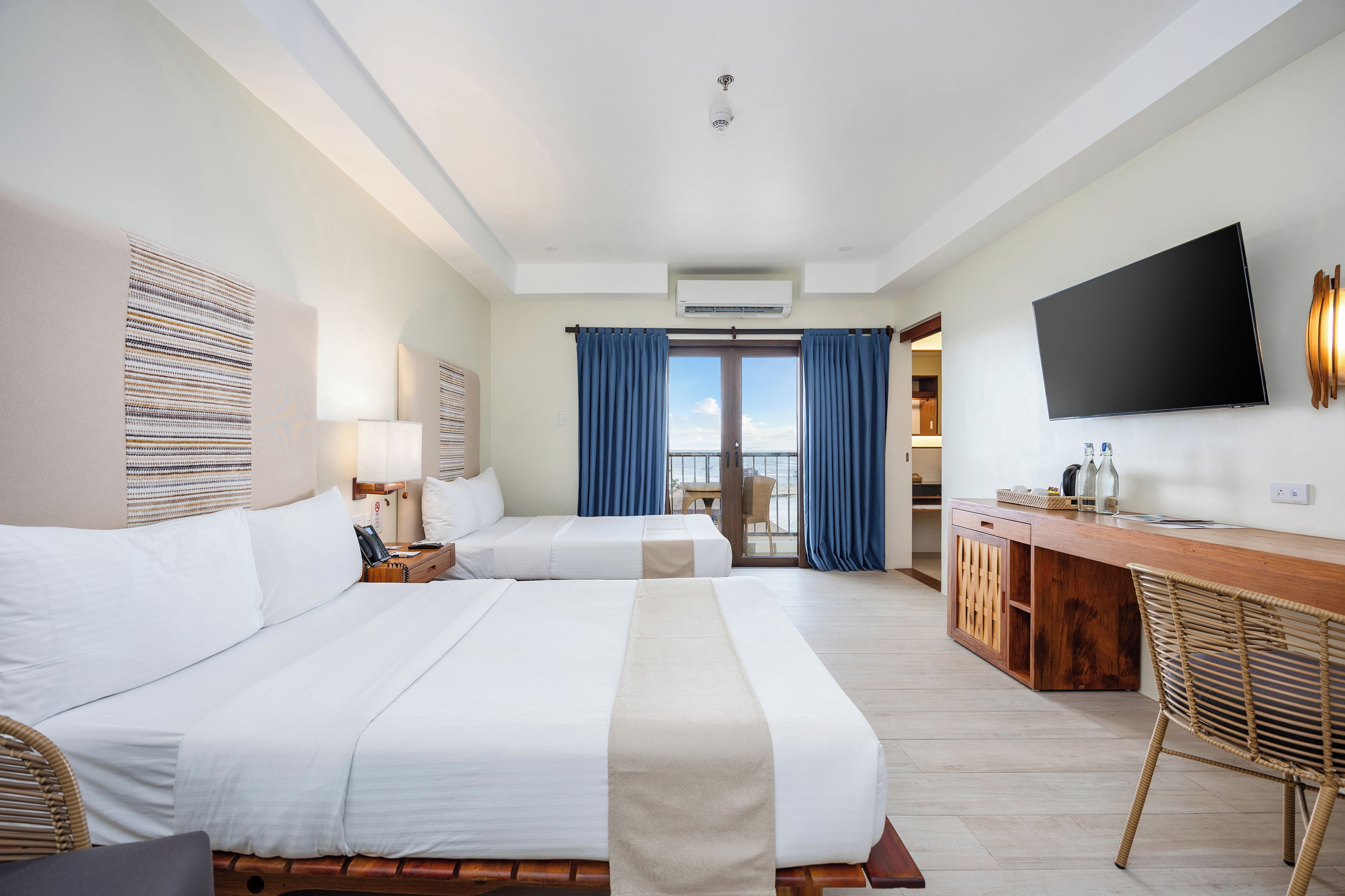 A hotel room with two beds, a desk, a flat-screen TV, and a balcony with curtains.
