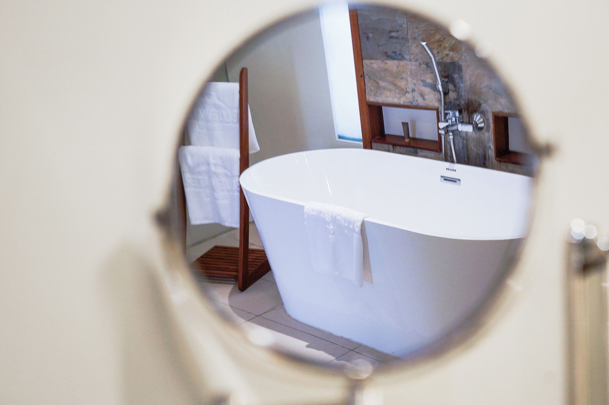 Un espejo redondo que refleja un baño con una bañera independiente, una silla de madera y un perchero.