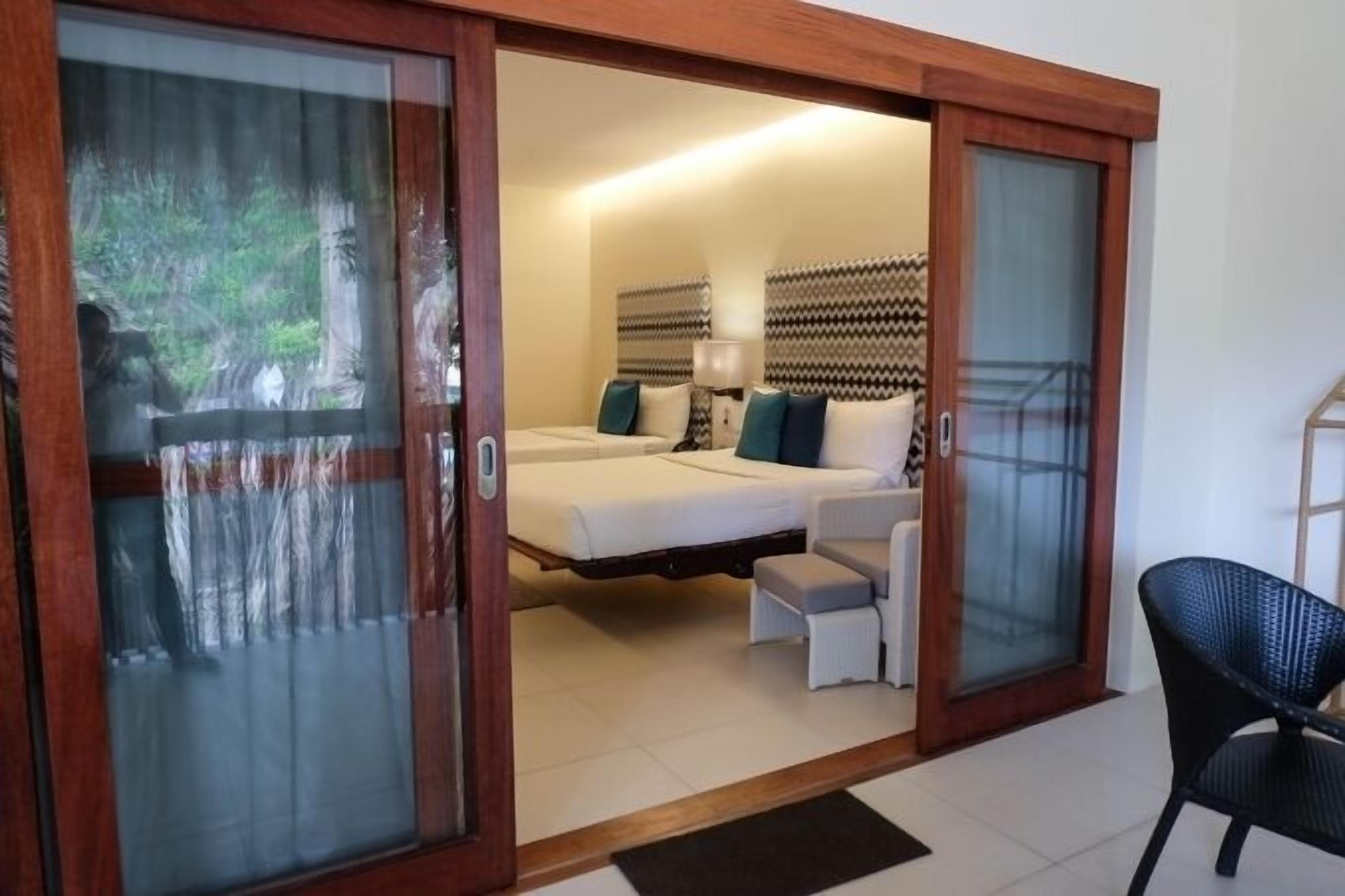 A hotel room with a sliding glass door leading to a balcony, a bed with pillows, a bedside table, and a chair.