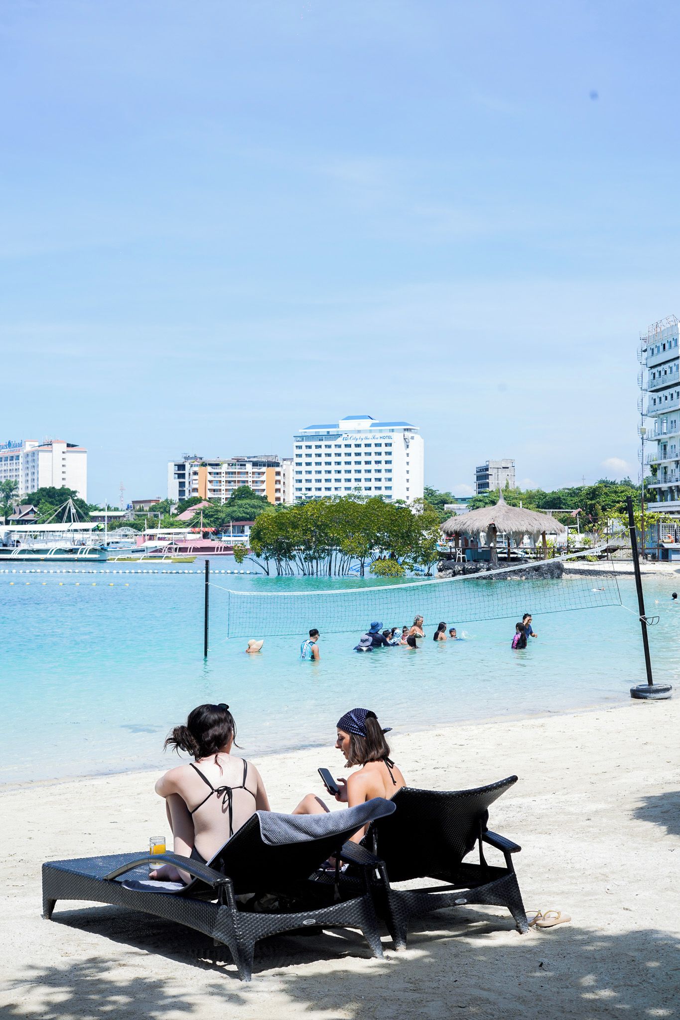 A beach with lounge chairs, a volleyball net, and a clear view of the sea and buildings in the background.