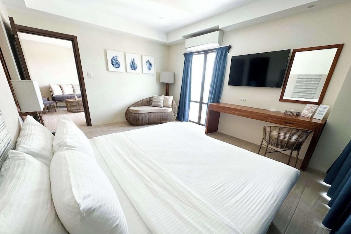 Habitación de hotel con una cama grande, un escritorio con silla, un televisor y una zona de estar con sofá y sillones.
