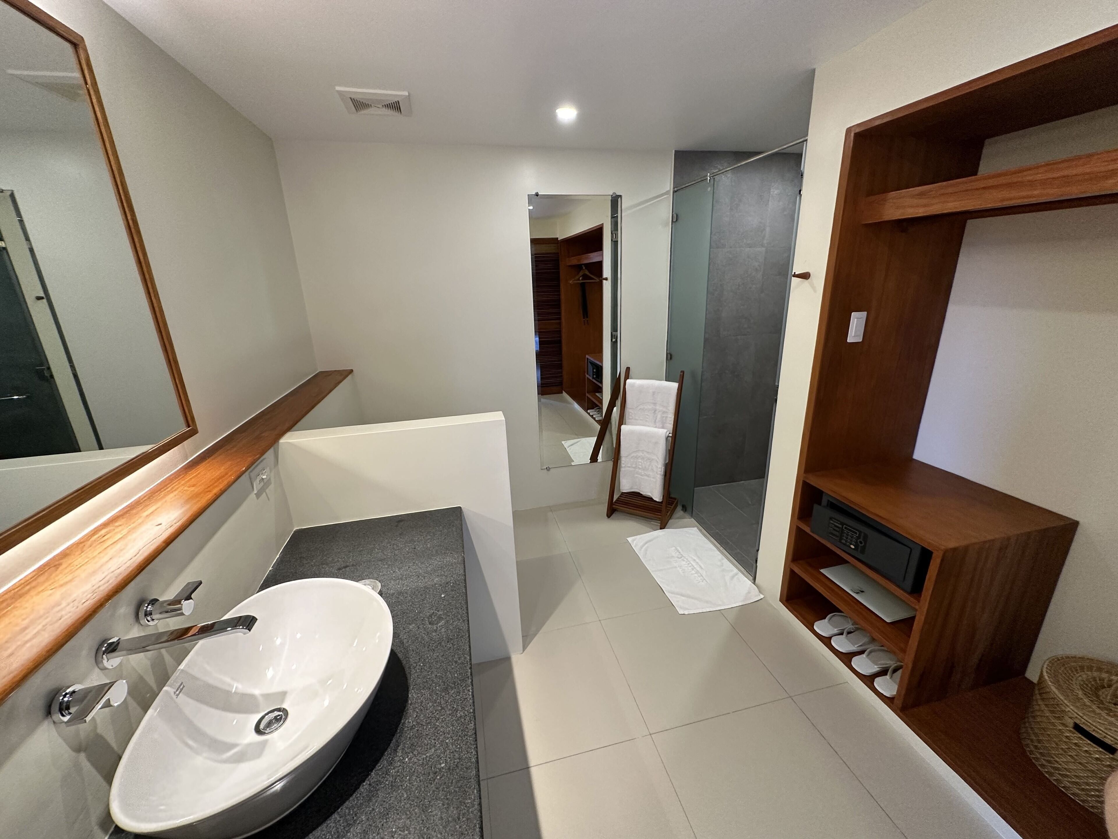 Een moderne badkamer met een witte wasbak, een houten wastafelmeubel, een glazen douchecabine en een houten plank met slippers.