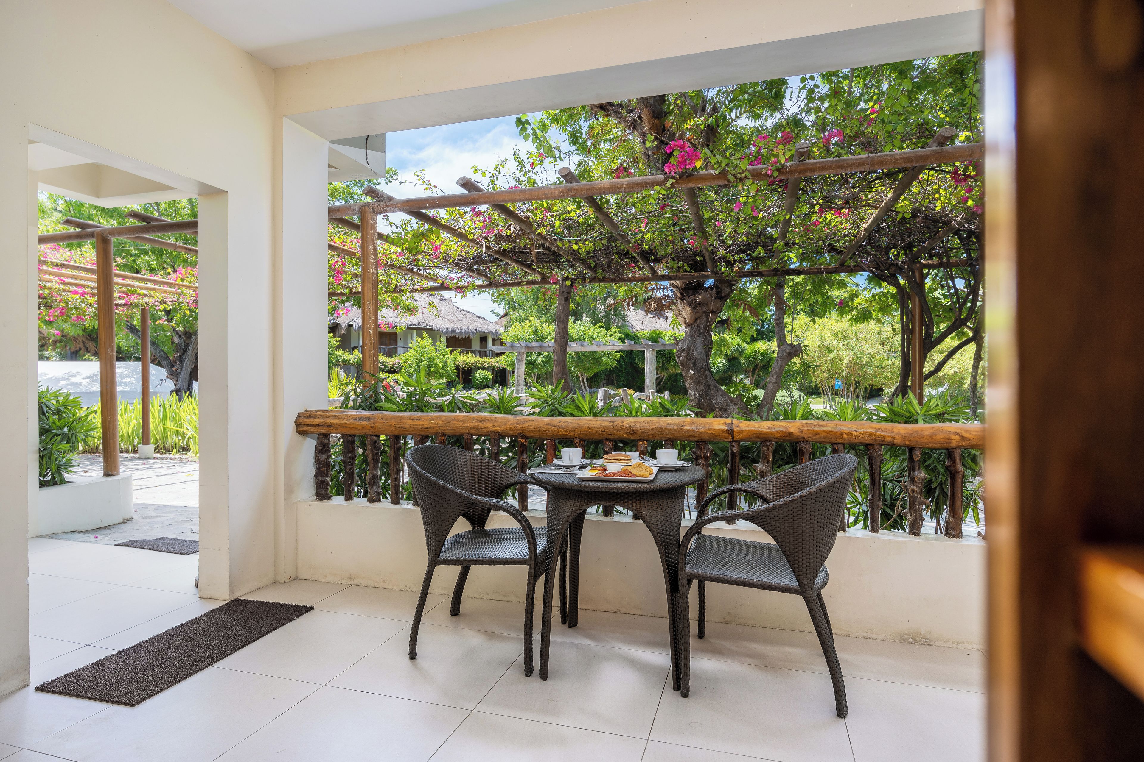 A balcony with a table and chairs, a pergola with climbing plants, and a view of a house in the background.