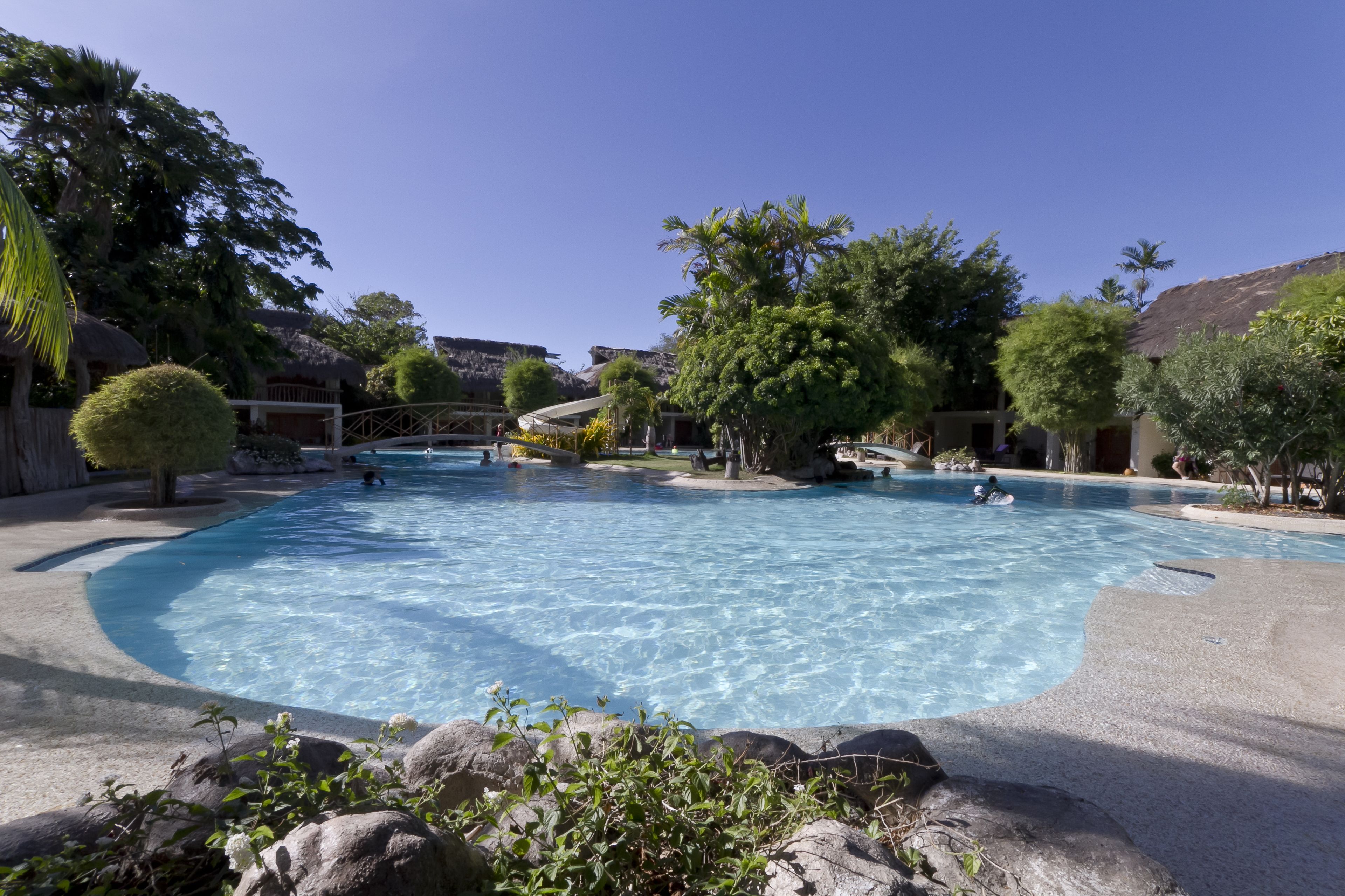 A large outdoor swimming pool surrounded by tropical vegetation and thatched-roof structures.