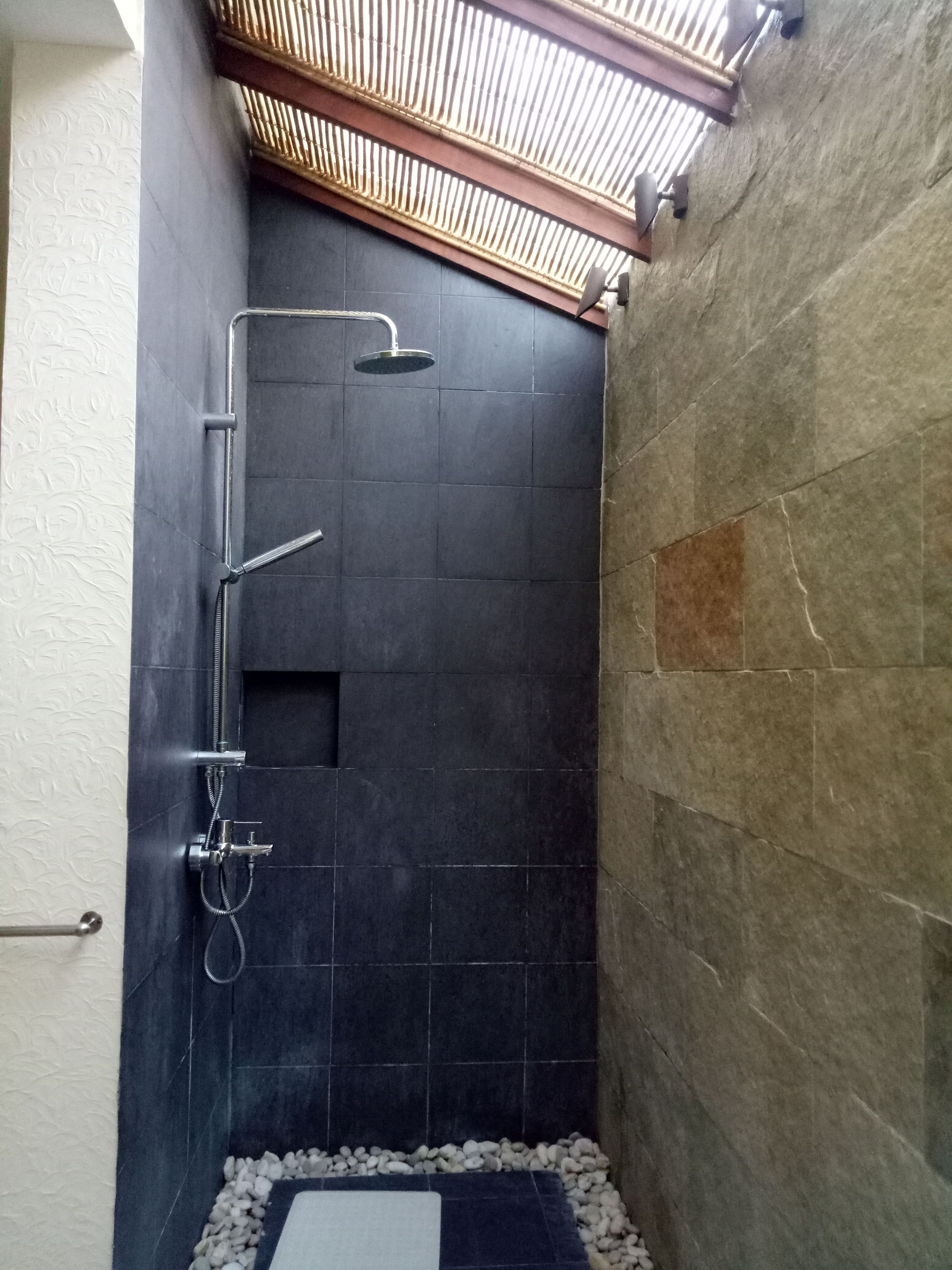 A small, dark-tiled shower area with a wooden ceiling and a white floor mat.