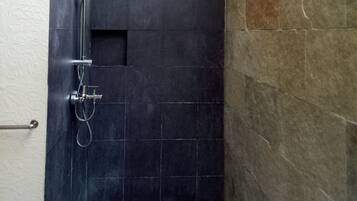 A small, dark-tiled shower area with a wooden ceiling and a white floor mat.