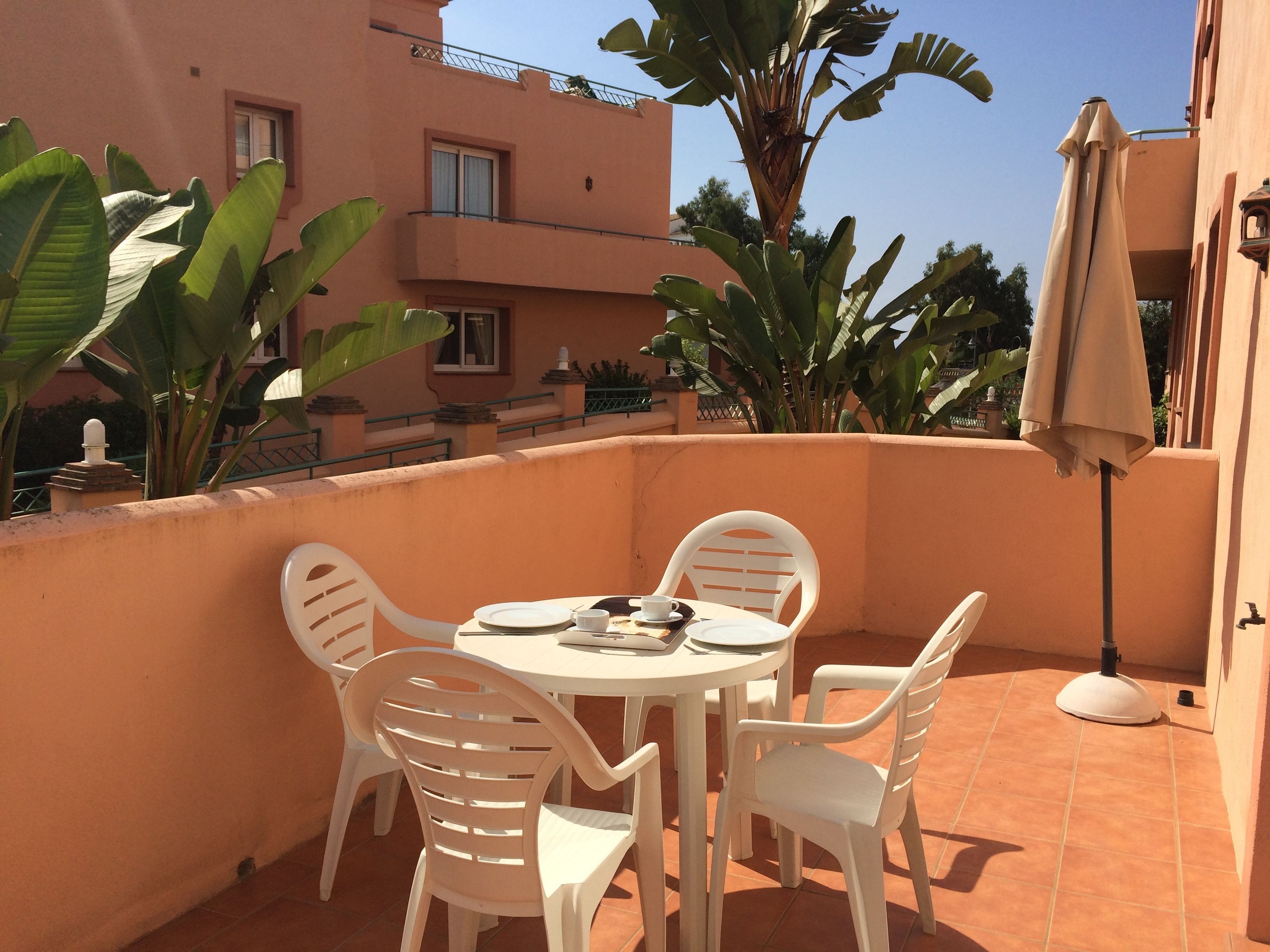 Un pequeño patio exterior con una mesa redonda y sillas de plástico blanco, rodeado por un muro naranja bajo y macetas con plantas.
