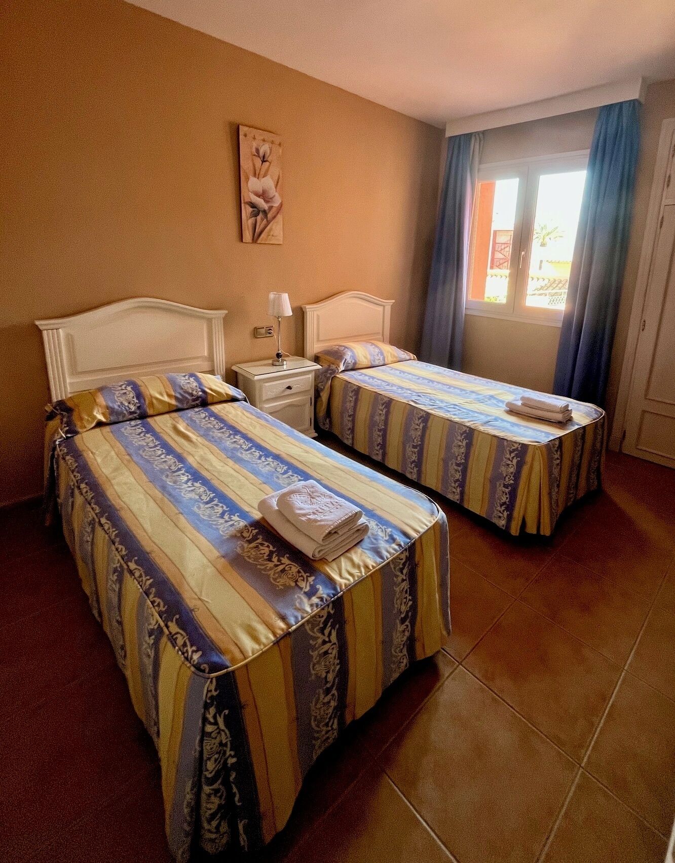 A hotel room with two beds, each with striped bedding, a nightstand with a lamp, a painting on the wall, and a window with curtains.