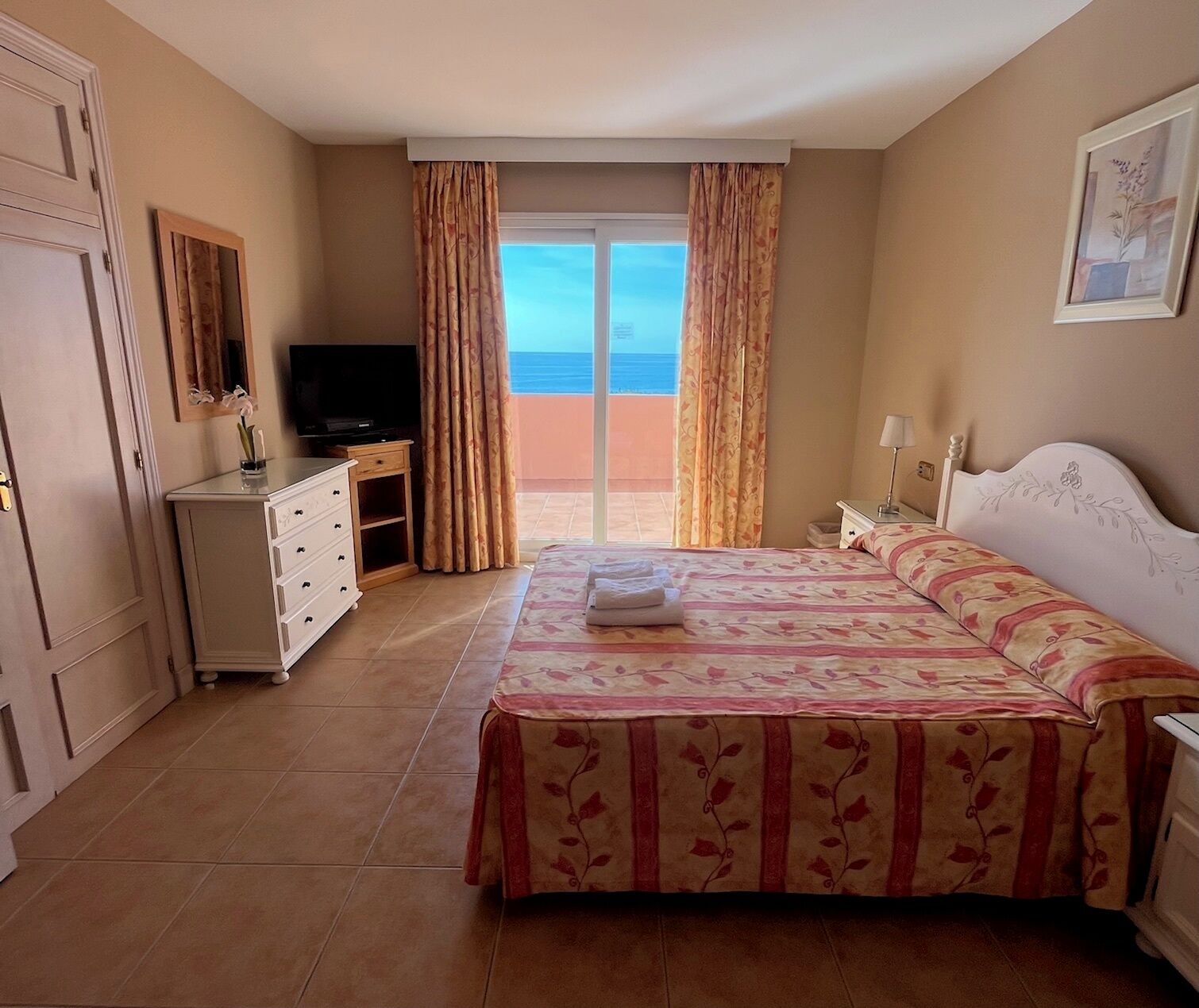 A hotel room with a bed, a TV, a desk, and a view of the sea.
