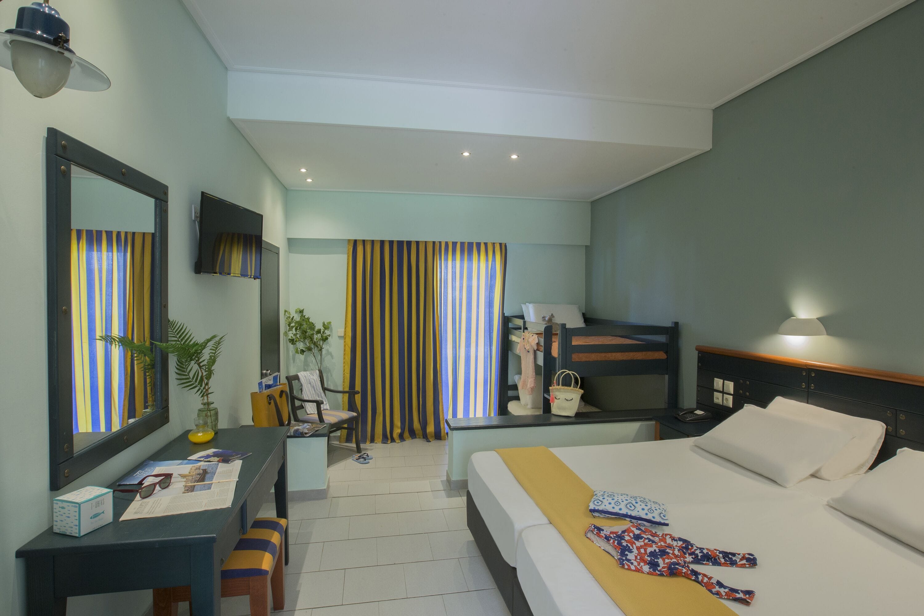 Une chambre d’hôtel comprenant un lit, un bureau avec une chaise, une table de chevet, un miroir, une lampe, une fenêtre avec des rideaux et une plante.