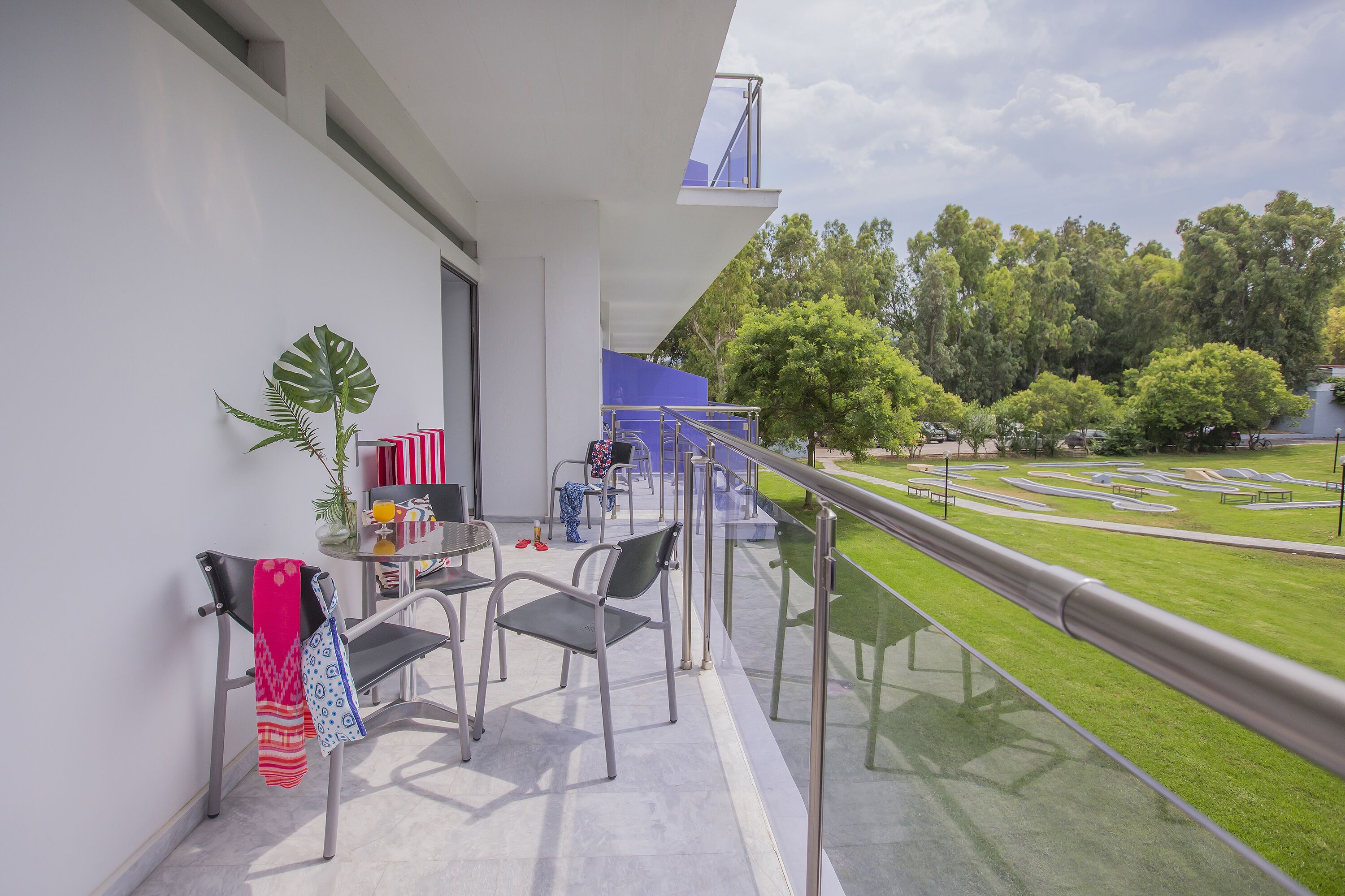 Uma varanda com guarda-corpo de vidro, uma mesa com cadeiras e vista para um gramado verde e árvores.