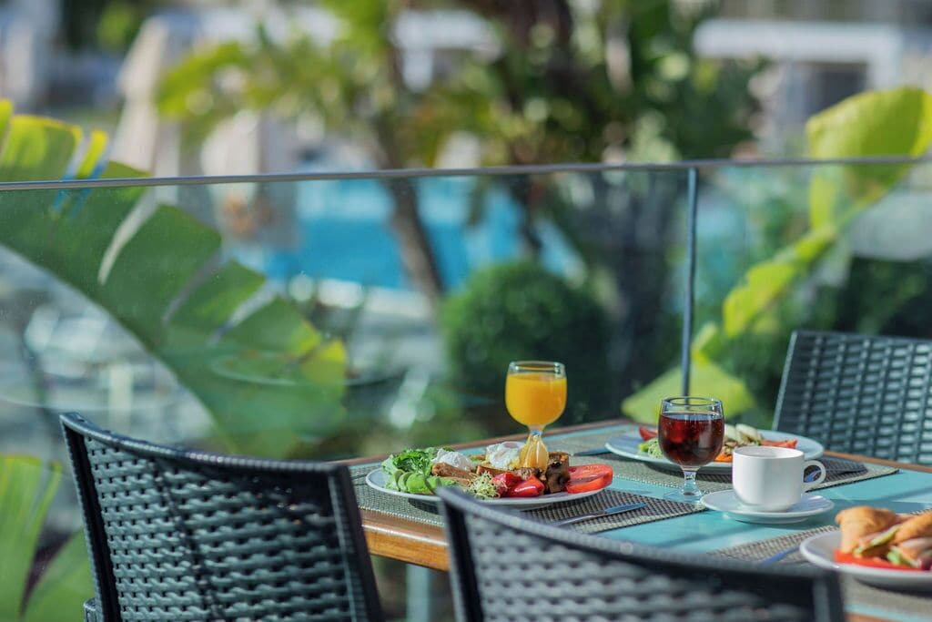 A dining table with plates of food, a glass of orange juice, and a glass of red wine, set against a backdrop of greenery and a pool.