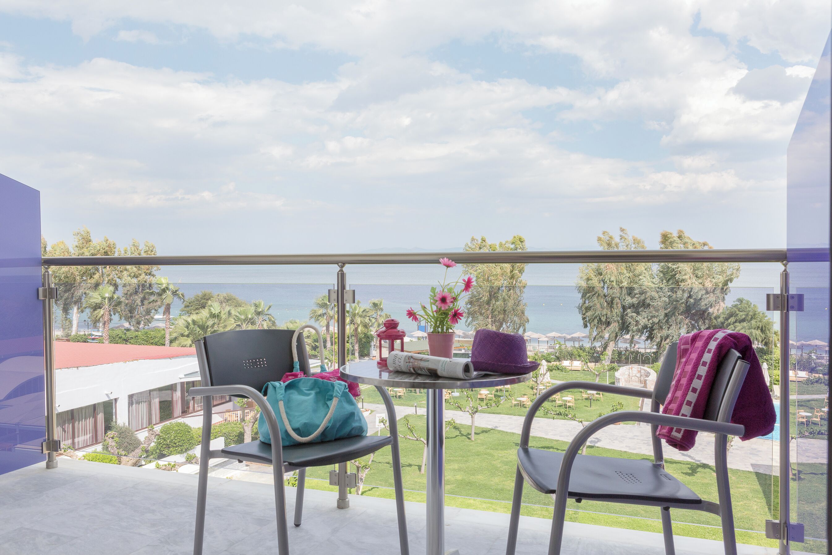 Un balcón con dos sillas, una mesa pequeña con un sombrero y un bolso, y vista al mar y al verdor.