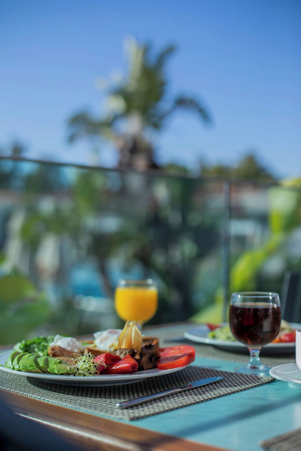 Une table à manger avec une assiette de nourriture, un verre de jus d’orange et un verre de vin rouge, le tout mis en valeur par un décor tropical.