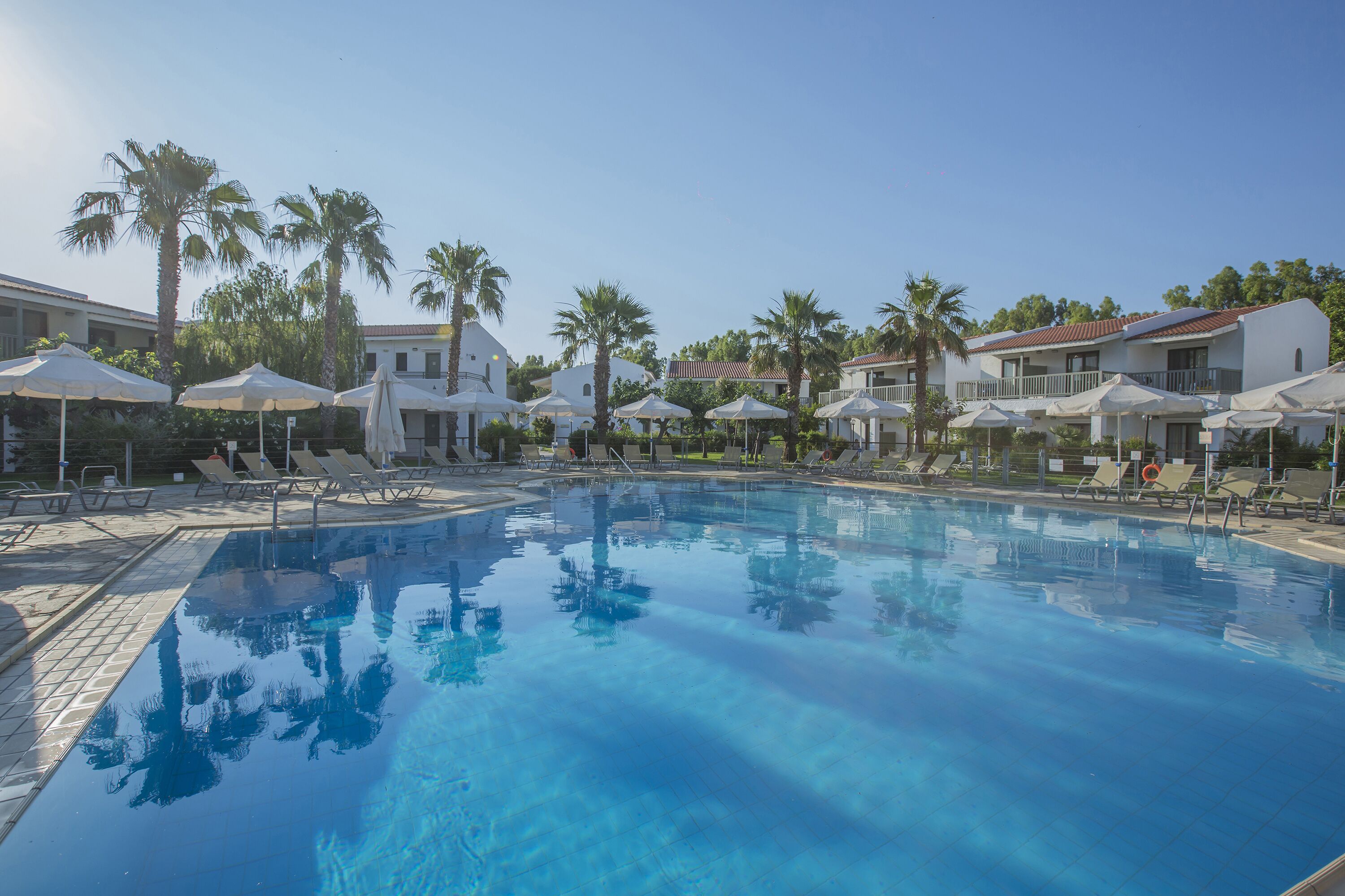 Ein Pool mit Liegestühlen und Sonnenschirmen, umgeben von Palmen und Gebäuden.