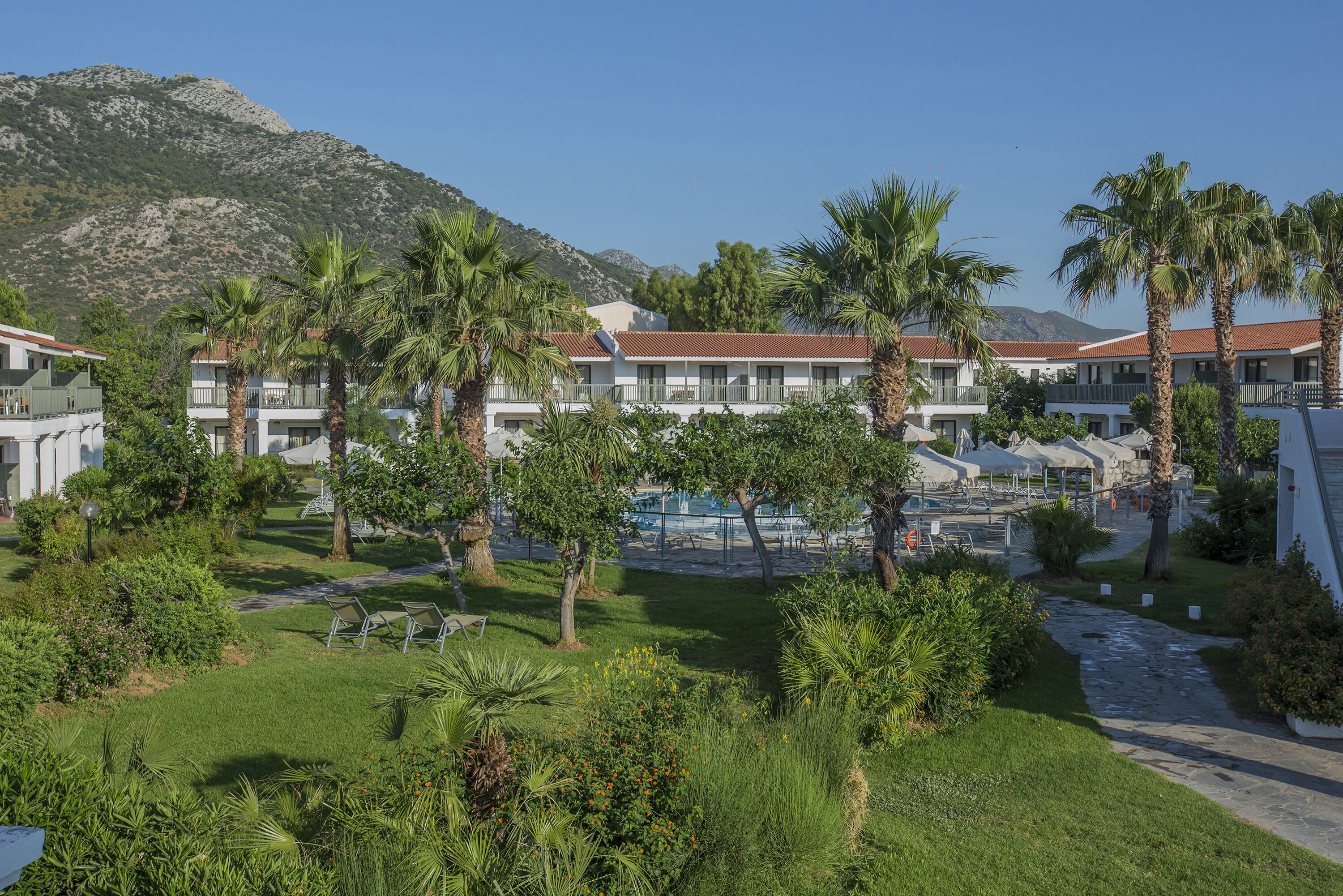 A resort with multiple buildings, palm trees, a pool area, and a mountainous backdrop.