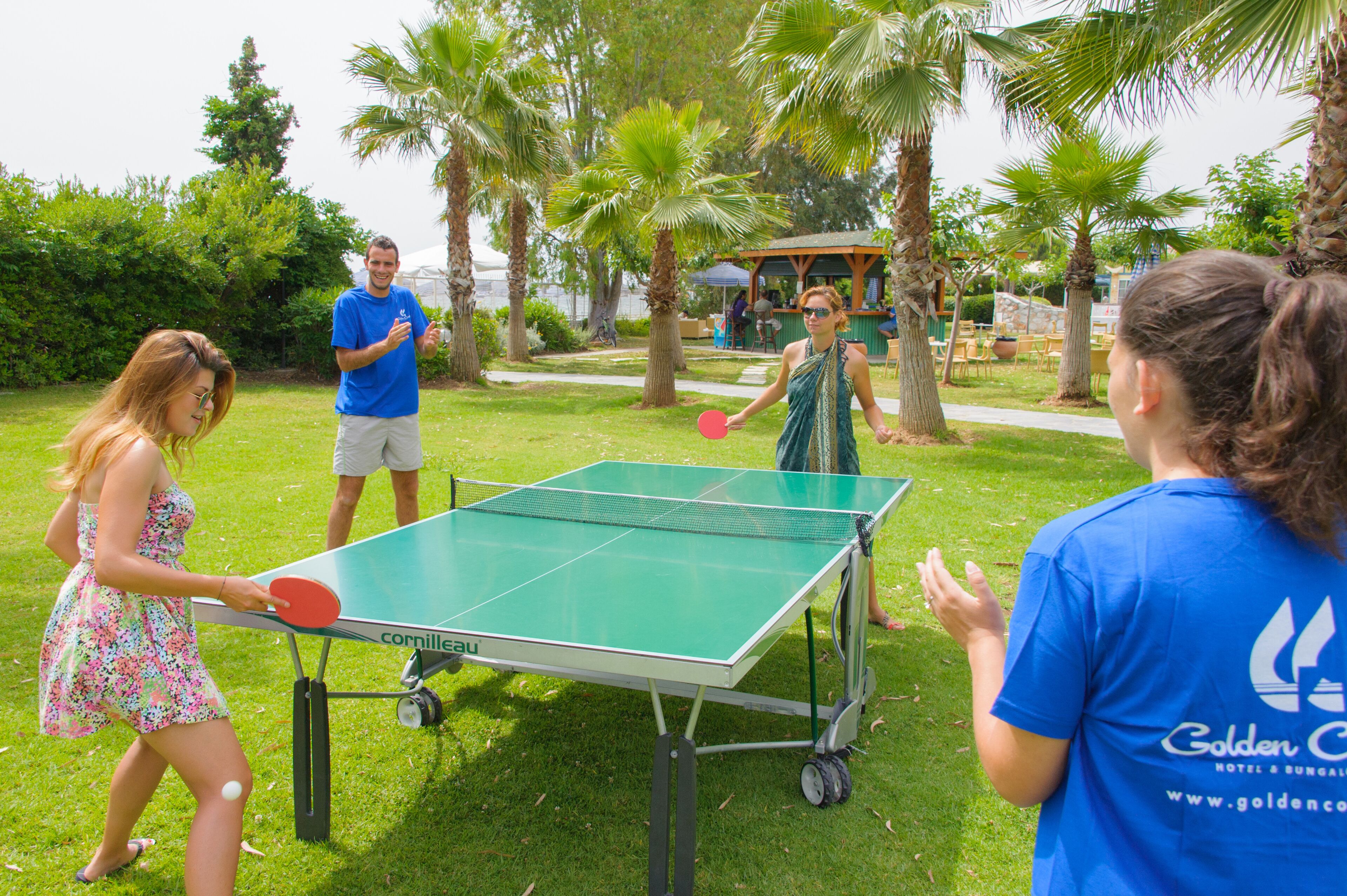 A table tennis table with a net, surrounded by people in a grassy outdoor area with palm trees.