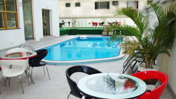 Una piscina con un tavolo e sedie in vetro in primo piano, un bar a bordo piscina a sinistra e un edificio con balconi sullo sfondo.