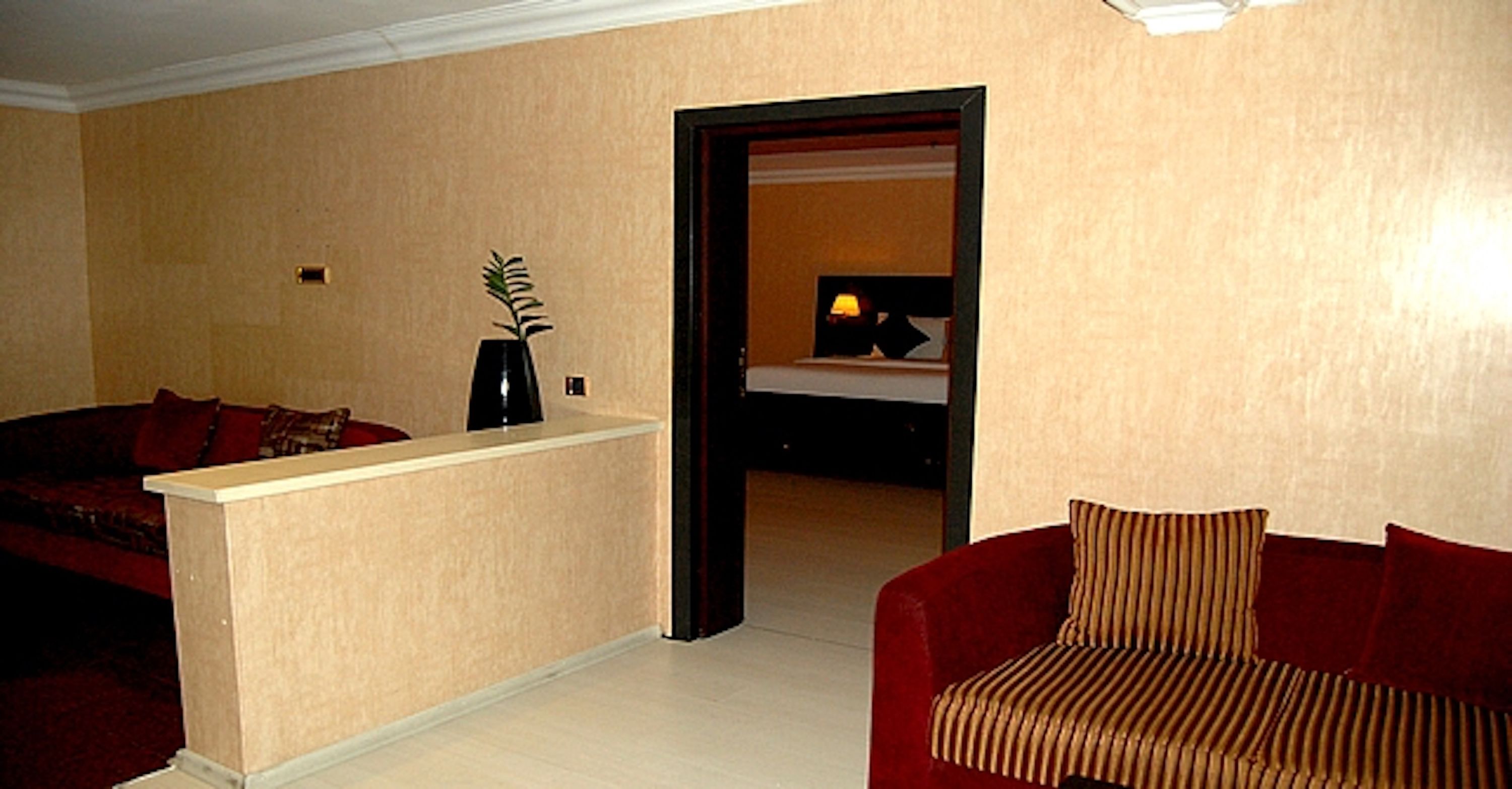 Een woonkamer met een beige bank, een rode bank, een kleine tafel met een vaas, en een doorgang naar een andere kamer.
