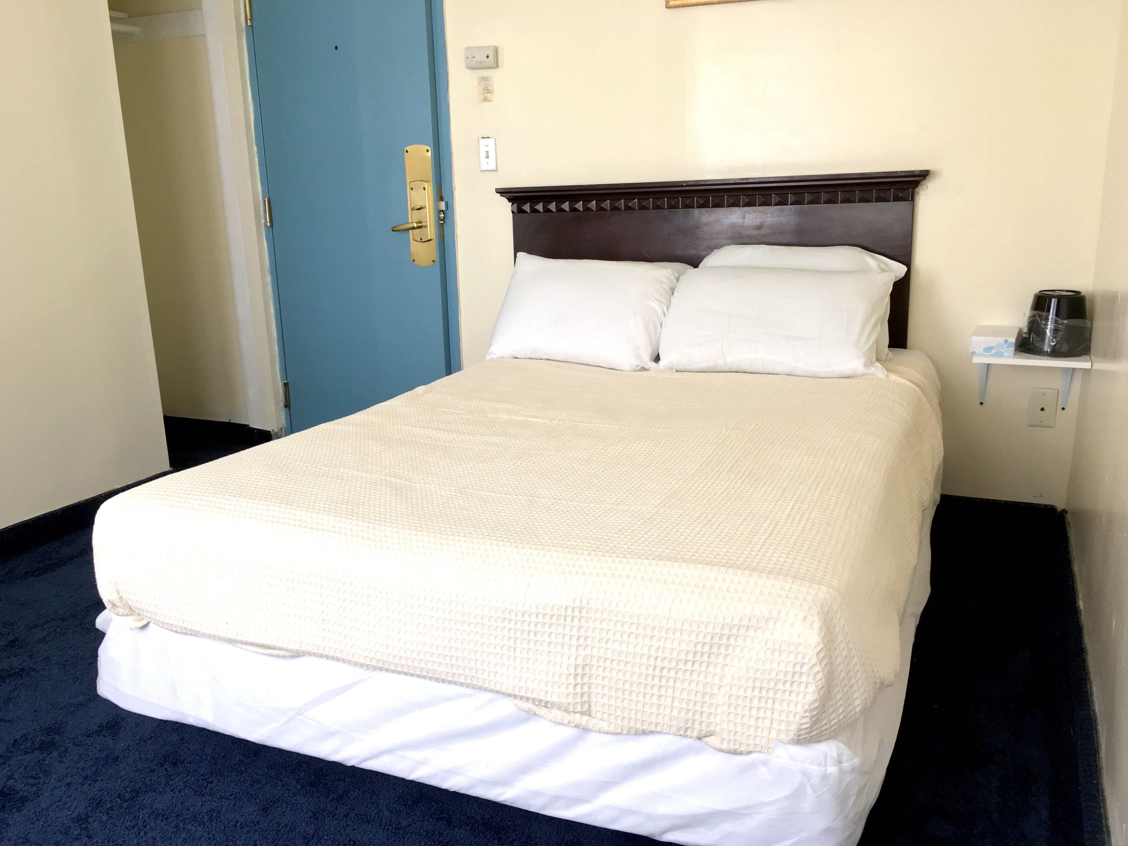 Una cama bien hecha con una colcha blanca y almohadas, un cabecero de madera, una mesita de noche con cafetera y una puerta azul al fondo.