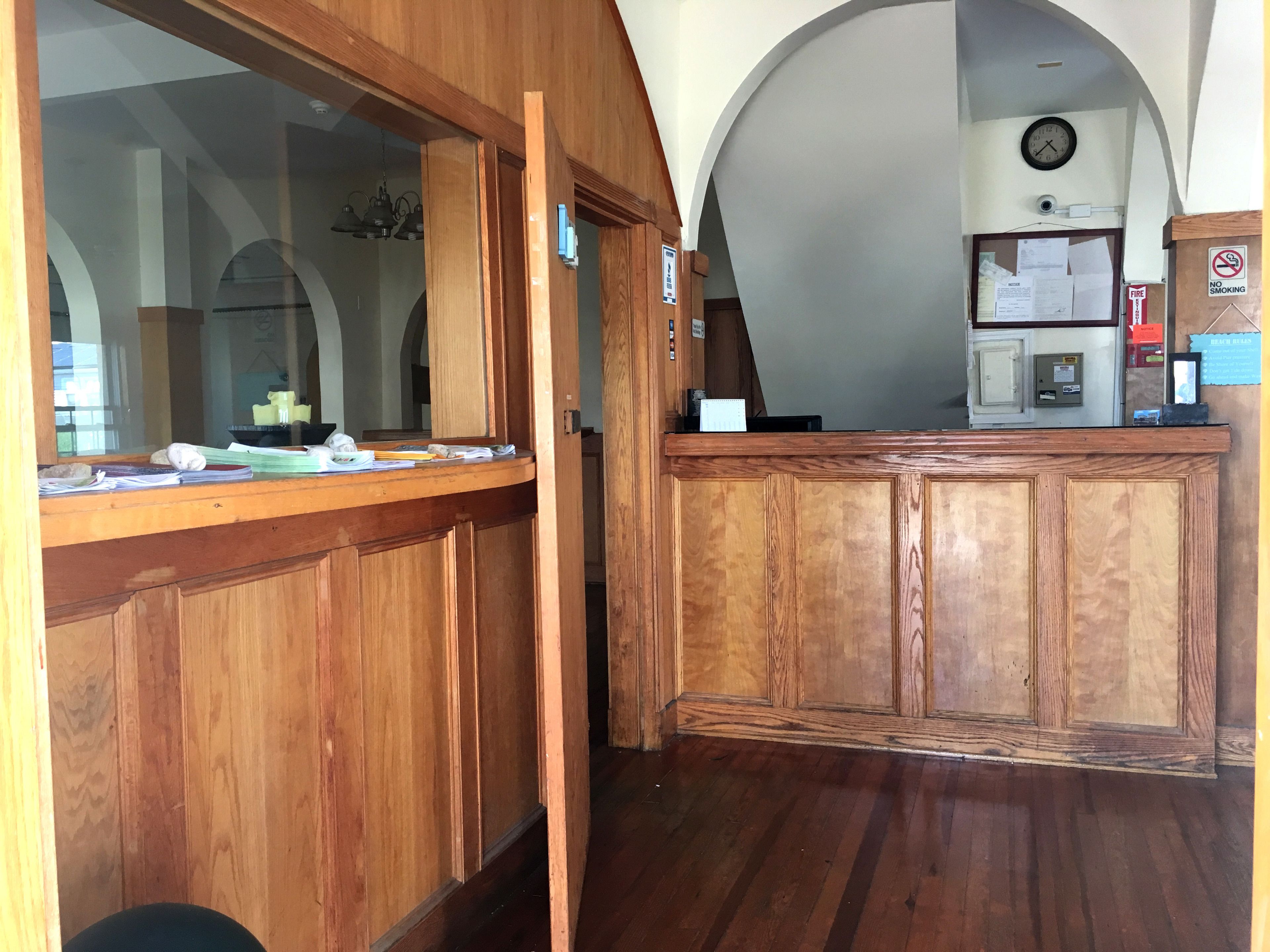A wooden reception desk with paperwork, a clock, and a no-smoking sign in a lobby with arched ceilings.