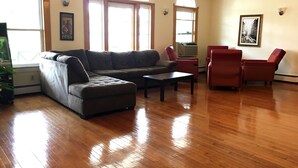 Una sala de estar con un sofá modular, una mesa de centro y dos sillones rojos. Hay ventanas grandes con arcos, un cuadro en la pared y un radiador.