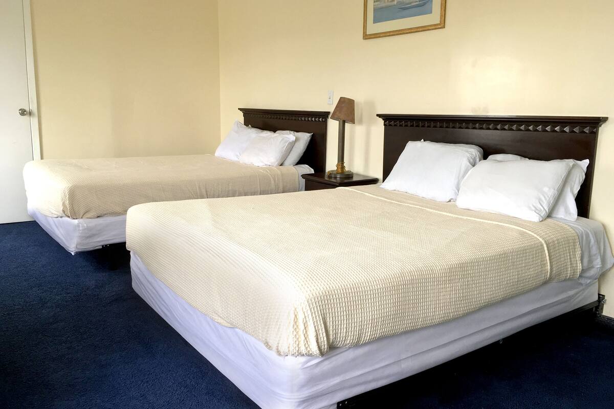 Dos camas individuales con ropa de cama blanca, una mesita de noche con una lámpara y un cuadro enmarcado en la pared.