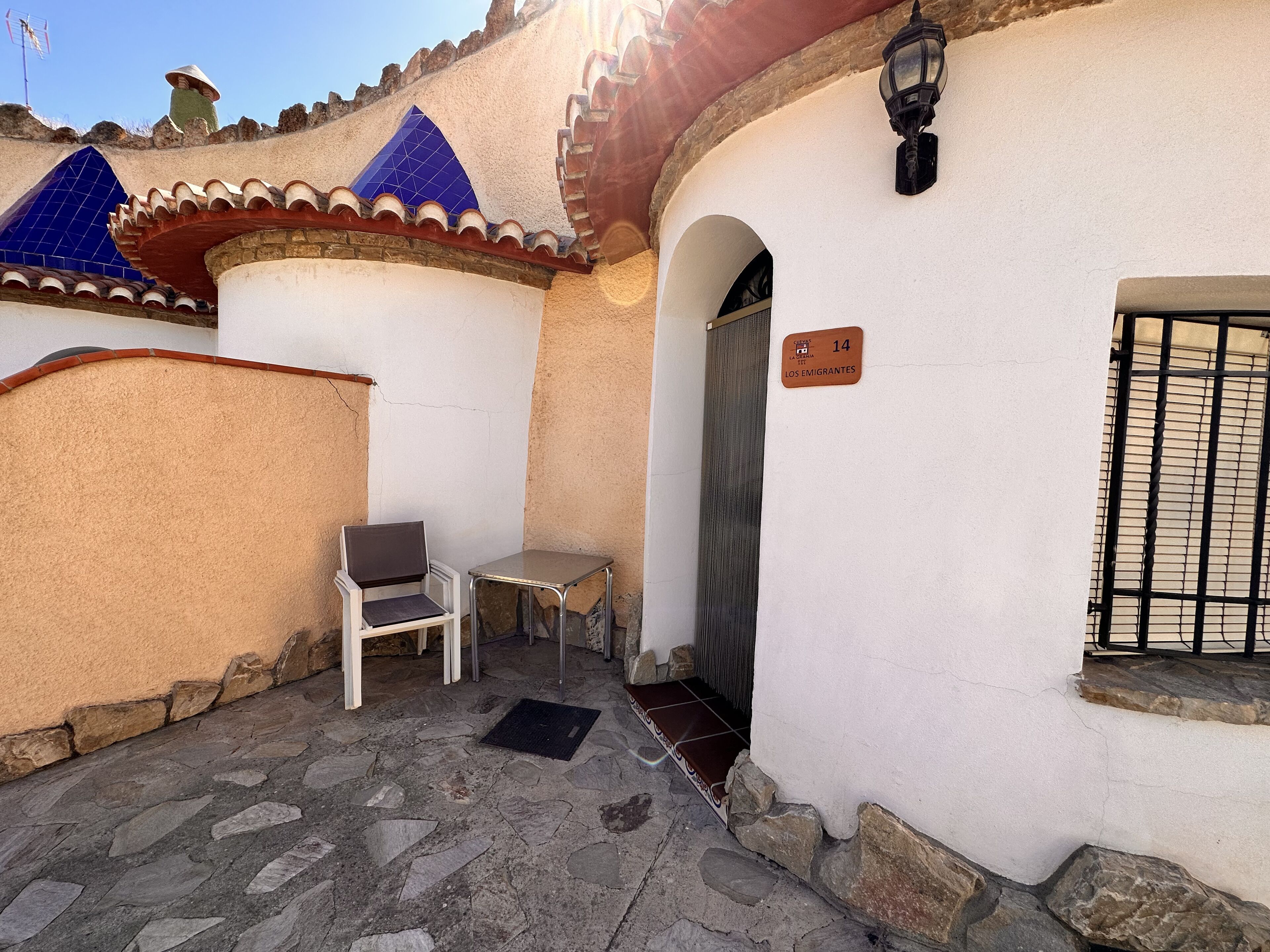 Une cour avec un sol en pierre, une chaise blanche, une petite table et une porte portant l’inscription « 14 Los Emigrantes ».