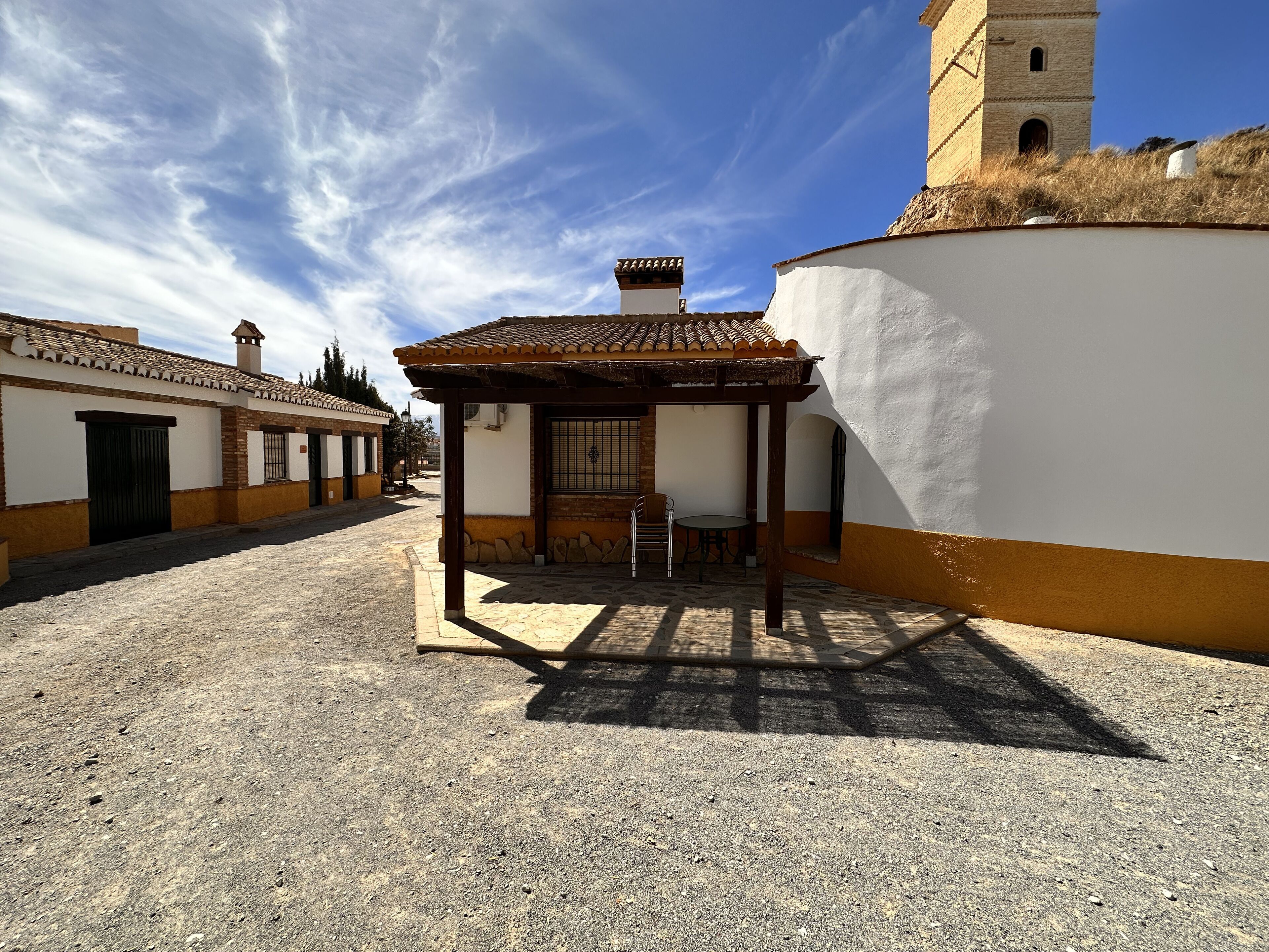 A courtyard with a covered seating area, a white building with a yellow base, and a tower in the background.