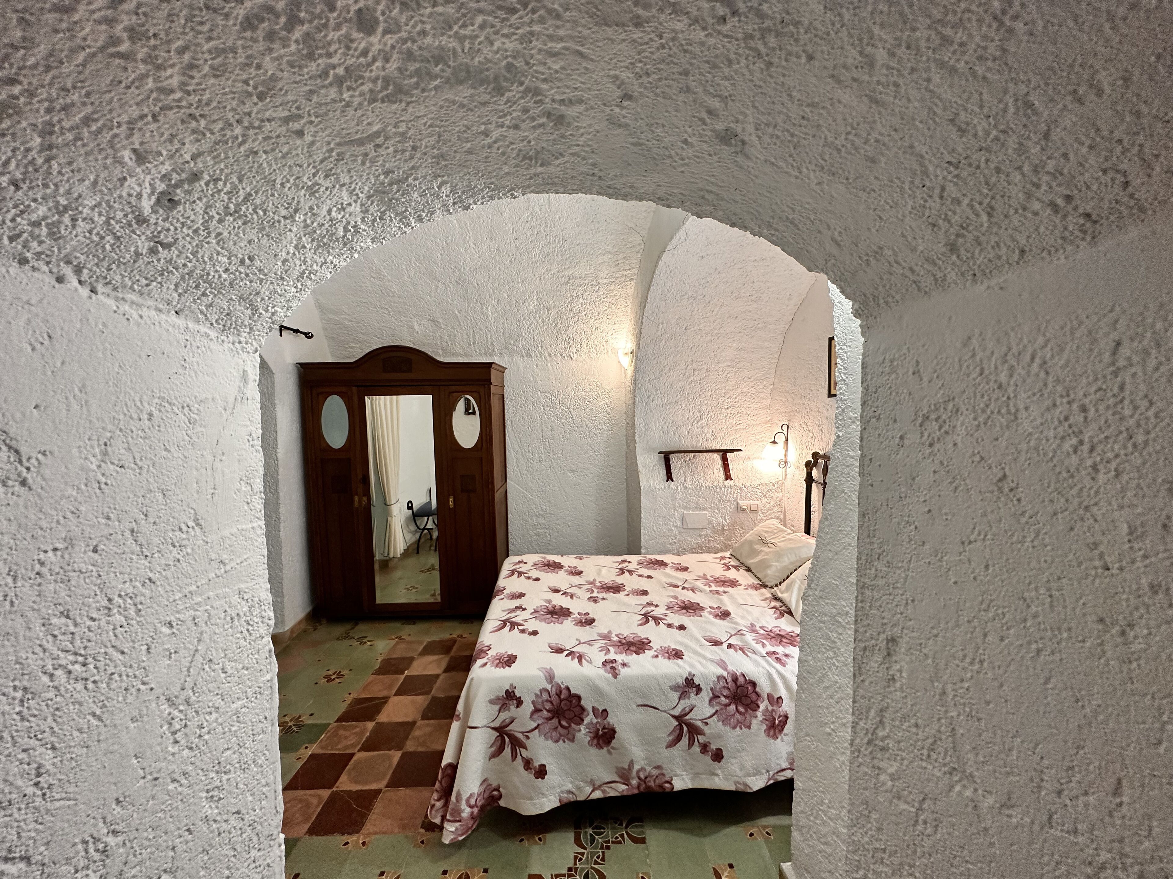 Ein Schlafzimmer mit Schachbrettmuster-Boden, einer blumigen Tagesdecke und einem Holzschrank mit Spiegel.
