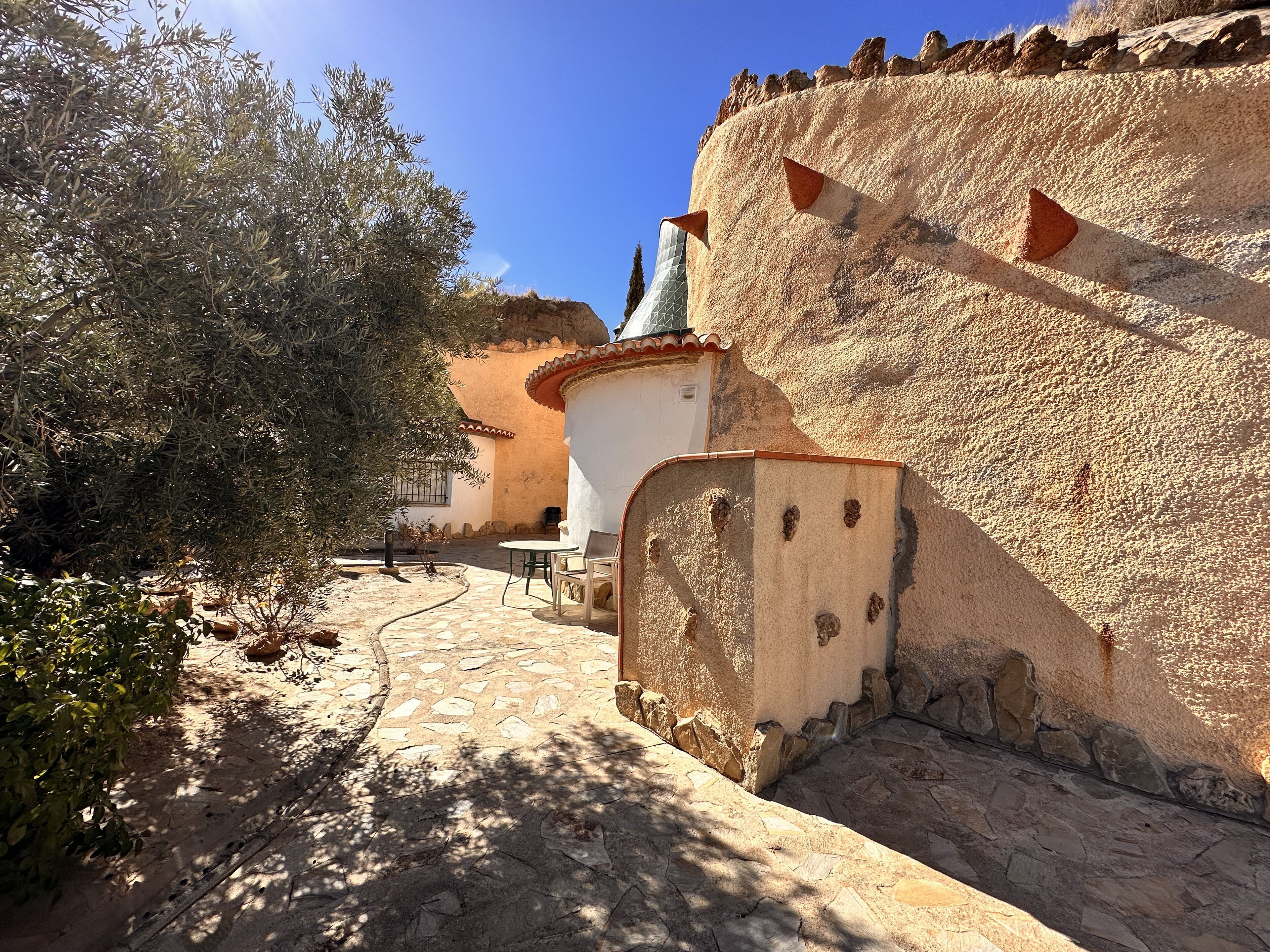 A courtyard with stone flooring, a white building with a red-tiled roof, and a wall with defensive features.