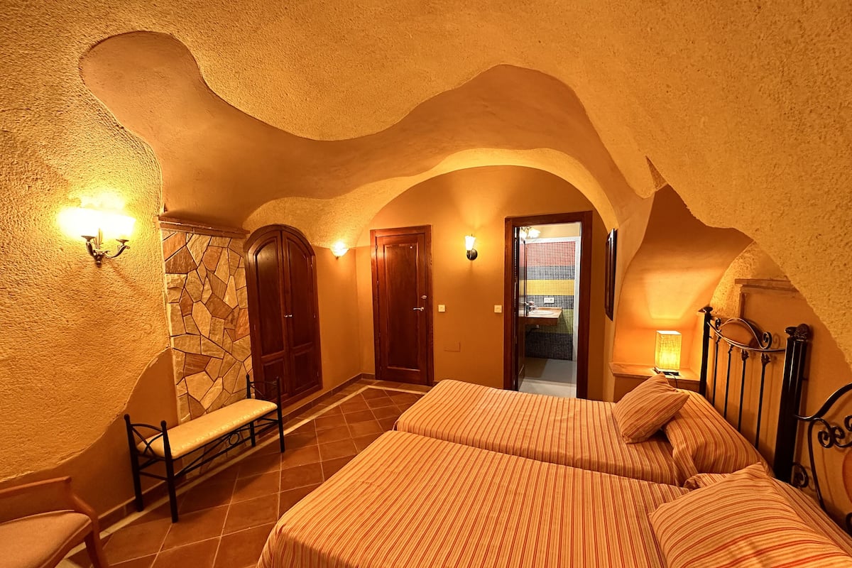 A bedroom with two beds, a bench, a stone wall, and a bathroom visible through an archway.