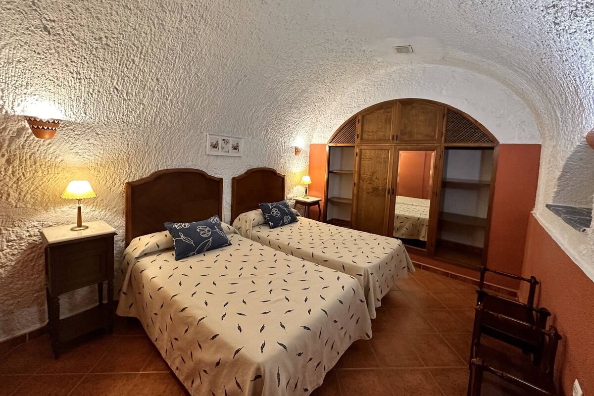 A bedroom with two beds, a wooden wardrobe, a small table with a lamp, and a tiled floor.