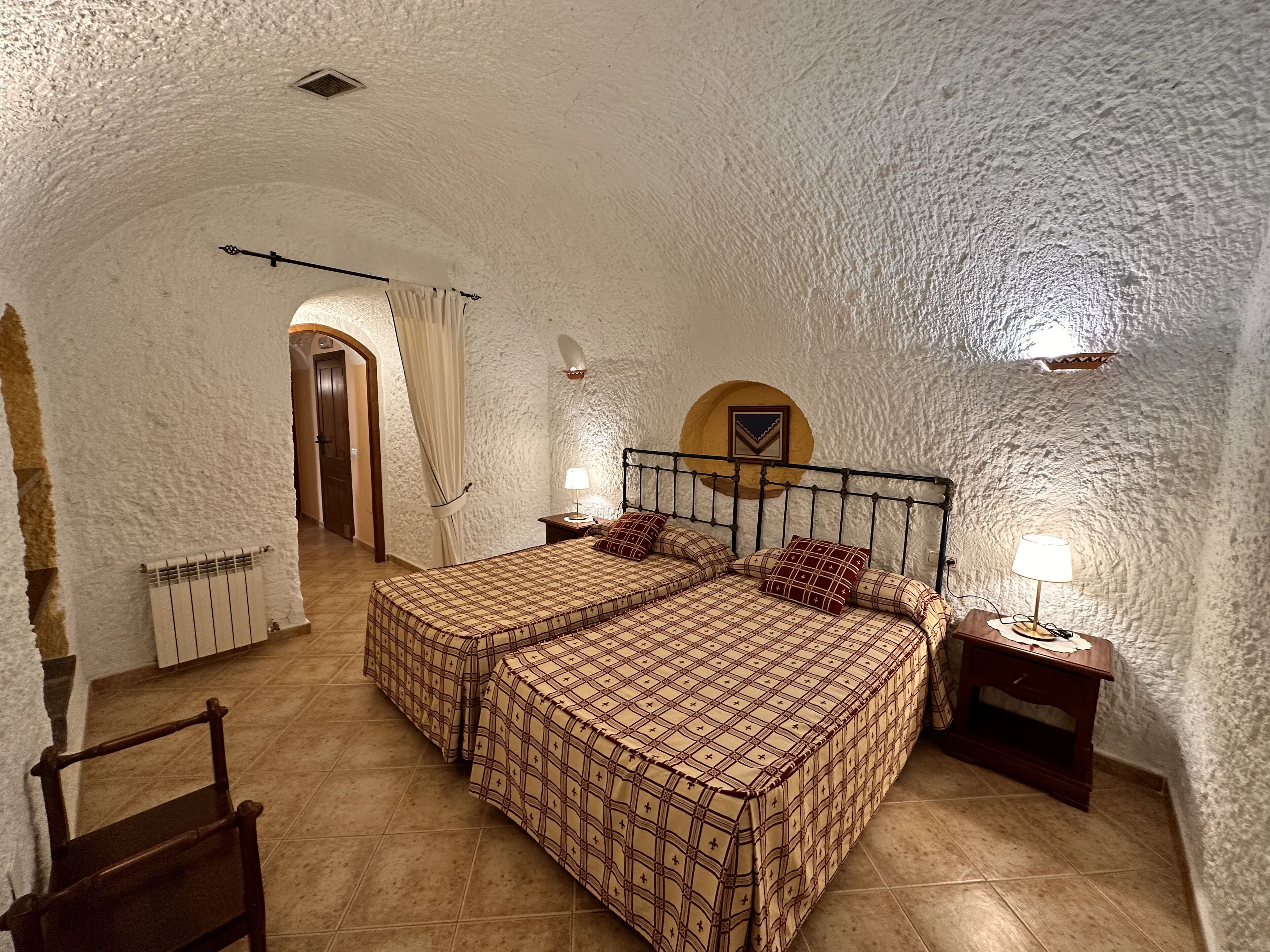 A cozy bedroom with two beds, a wooden chair, a nightstand with a lamp, and a rustic stone wall.