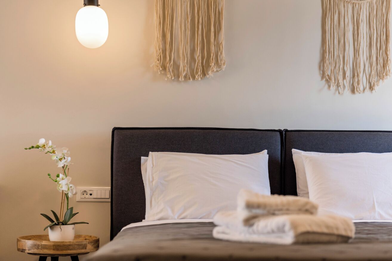 A neatly made bed with white pillows and a folded blanket, a bedside table with a potted orchid, and macramé wall hangings.