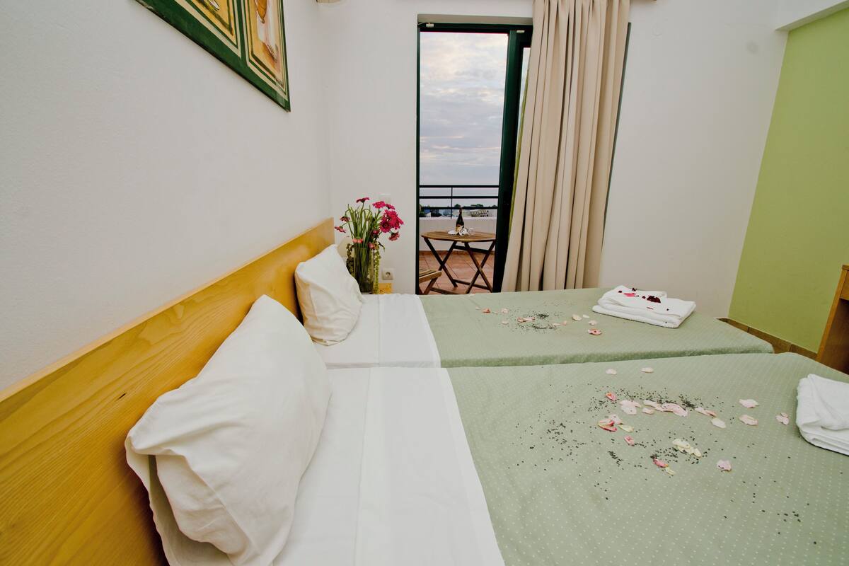 A hotel room with a bed, a small table with a vase of flowers, a view of the outside balcony, and a door leading to the balcony.