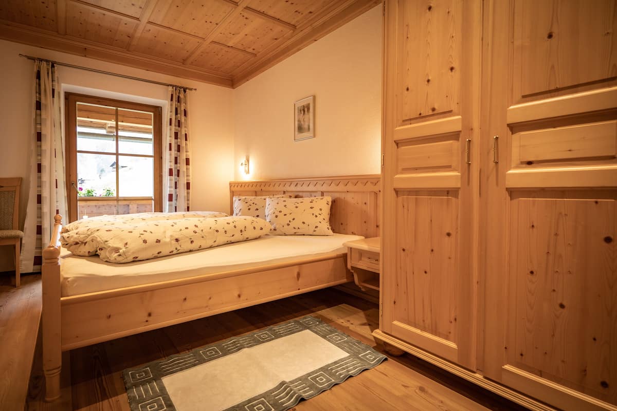 A wooden bed with a headboard, a bedside table, a nightstand, a window with curtains, and a wooden wardrobe.