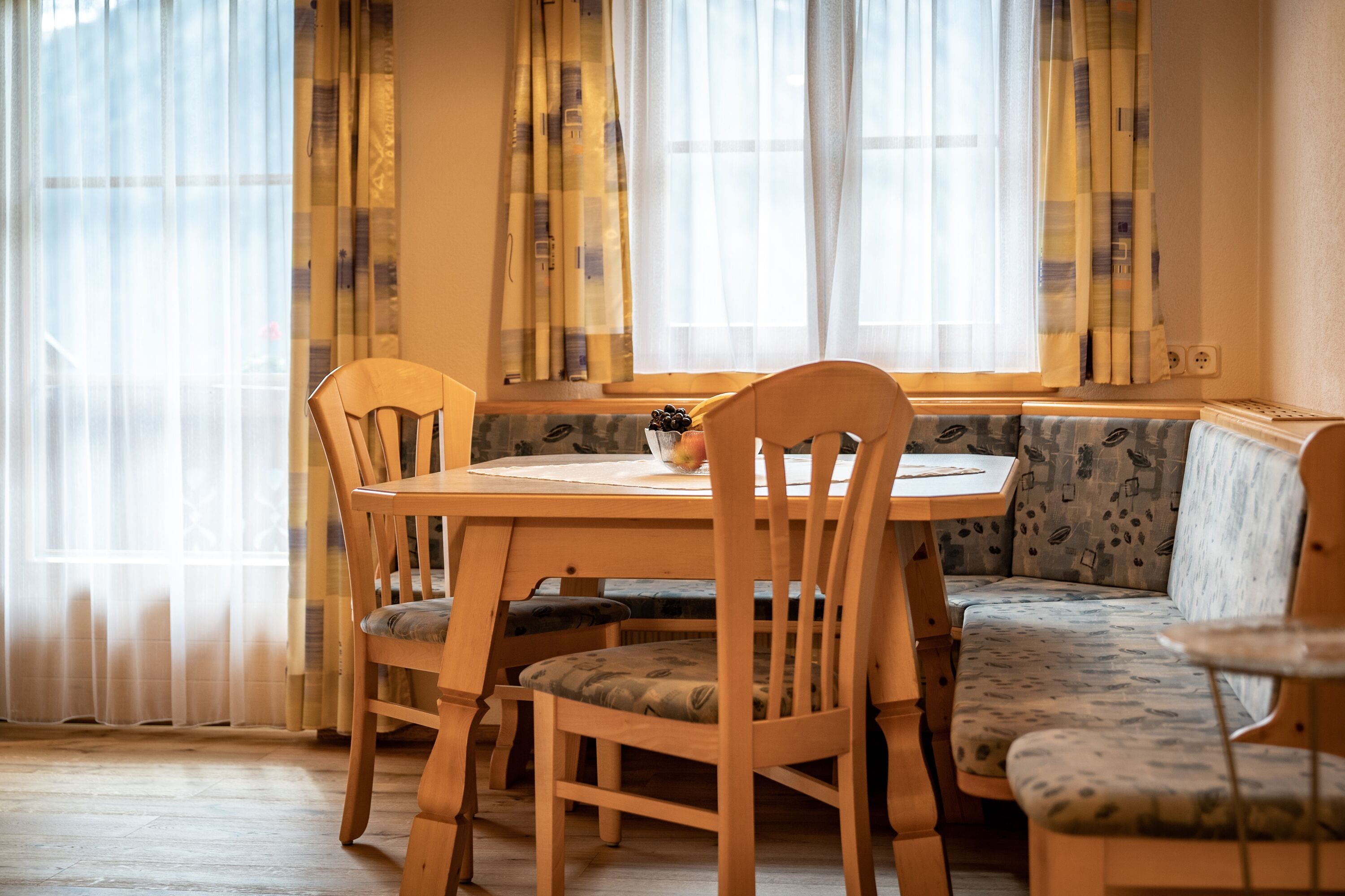 A dining area with a wooden table and chairs, a bench with patterned upholstery, and a window with sheer curtains.