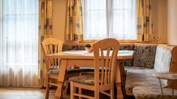 A dining area with a wooden table and chairs, a bench with patterned upholstery, and a window with sheer curtains.