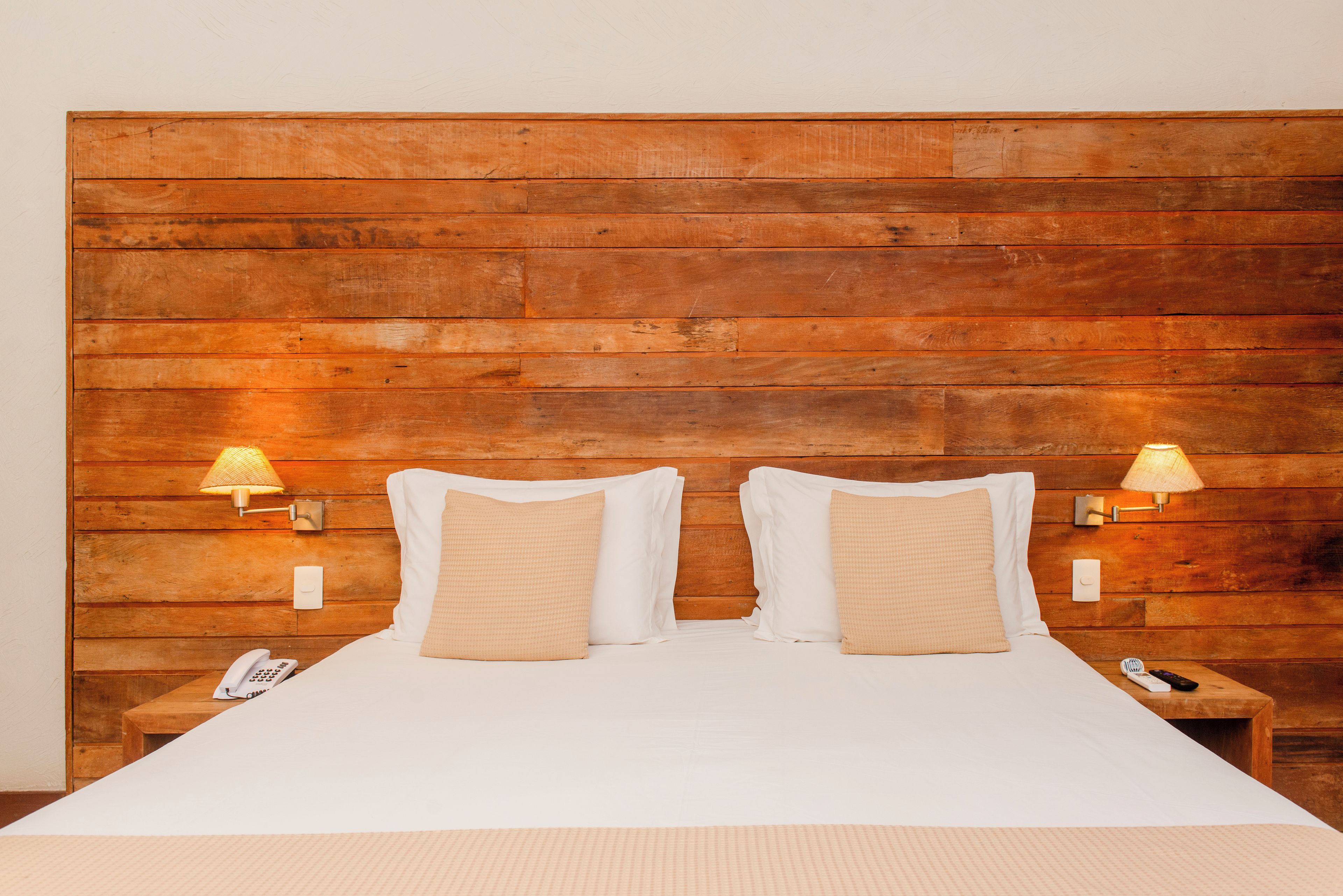 A hotel room with a wooden headboard, two bedside lamps, a bed with white linens, and a bedside table with a phone and remote control.