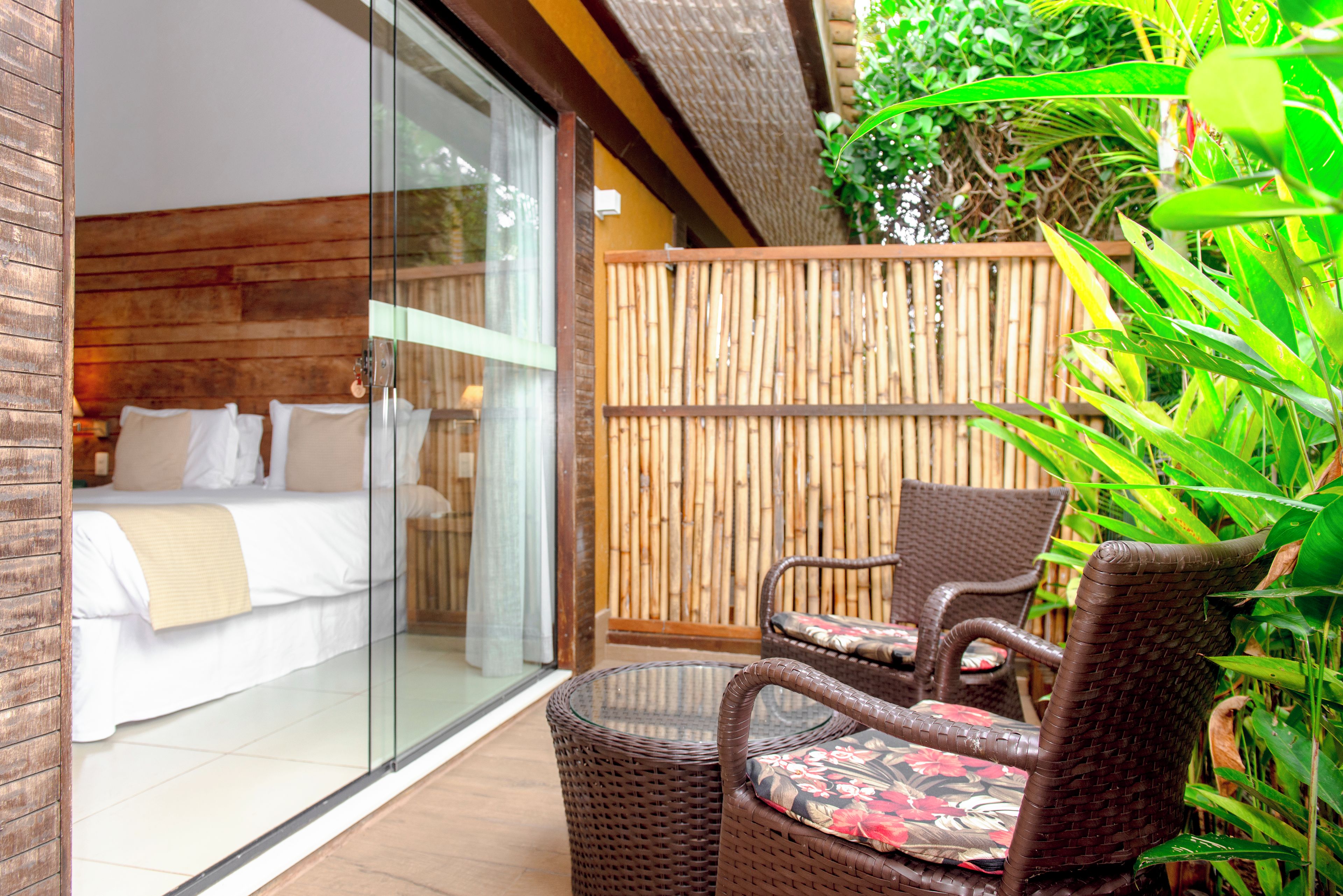 A room with a bed, a glass door leading to a balcony, wicker furniture, and a bamboo fence.