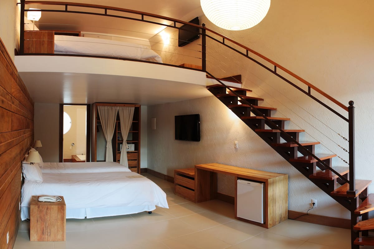 A bedroom with a bunk bed, a desk, a chair, a small refrigerator, a wooden staircase, and a television.